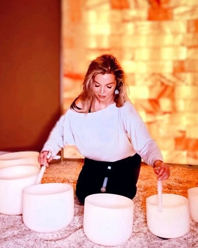 A woman is sitting on the floor, playing crystal singing bowls with a wooden mallet in a cozy indoor space with a warm, textured wall in the background.