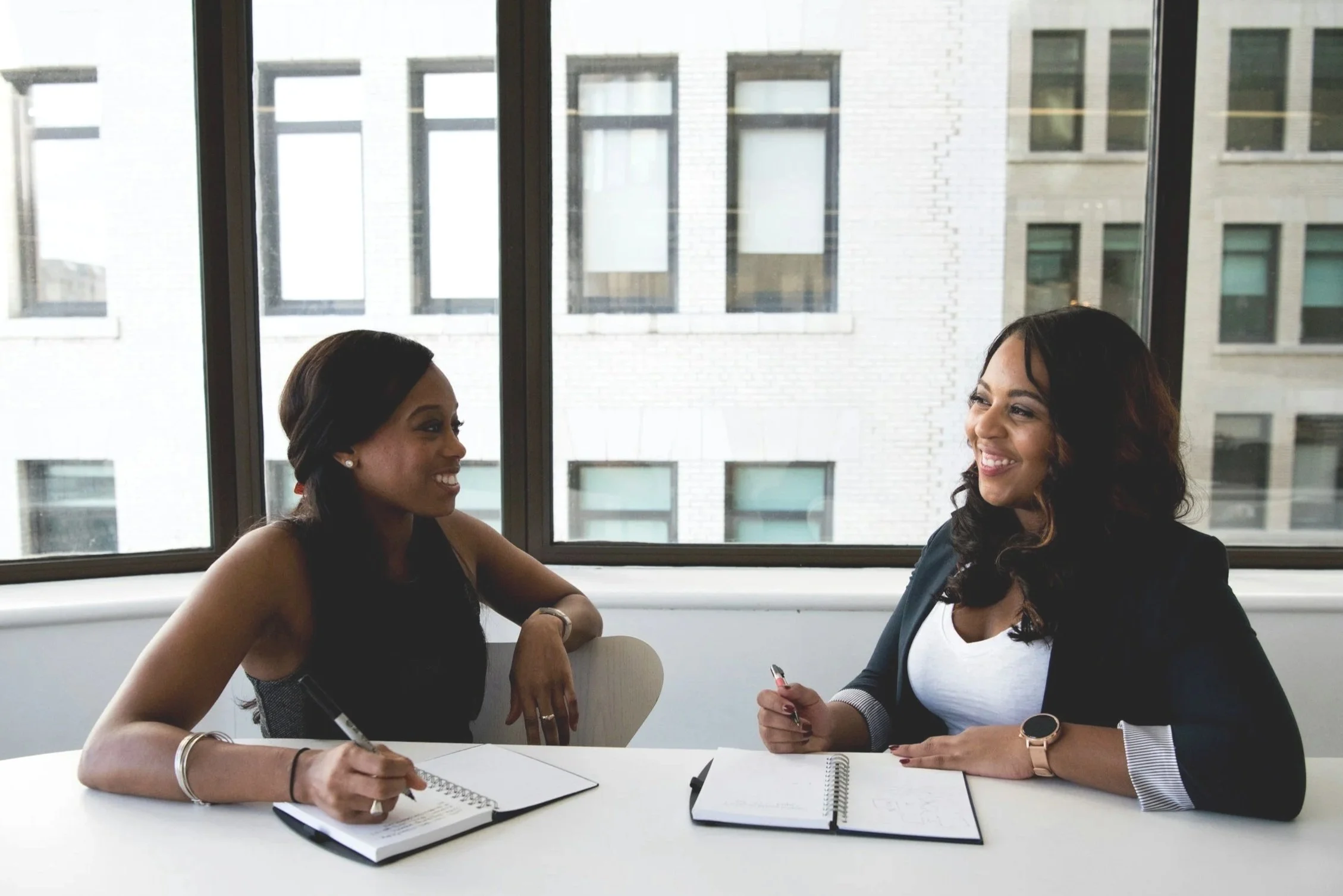 Two women are sitting at a table having a friendly conversation, with notebooks and pens in front of them, near a large window showing neighboring buildings.