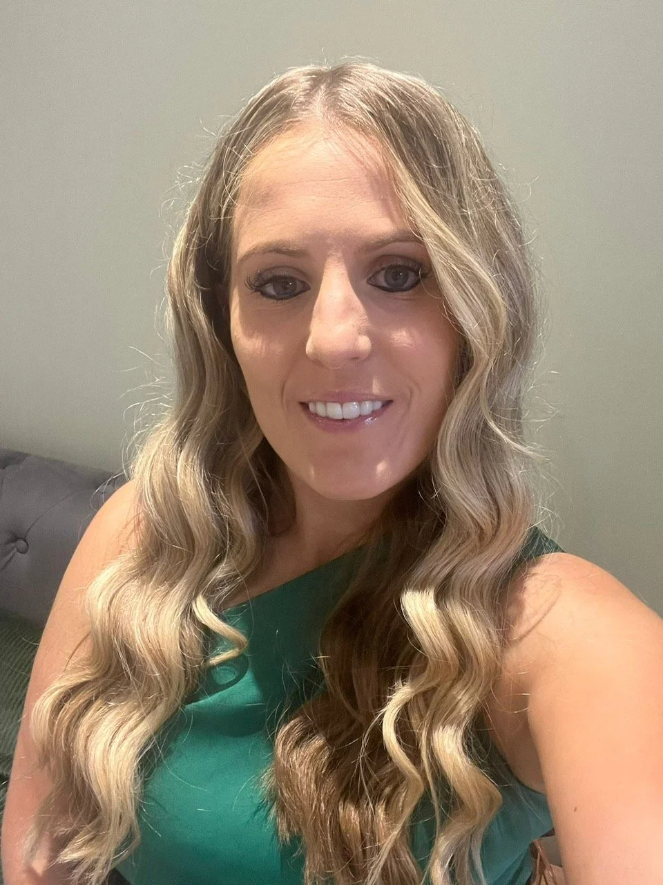 Natasha, a woman with wavy blonde hair, is taking a selfie and smiling.