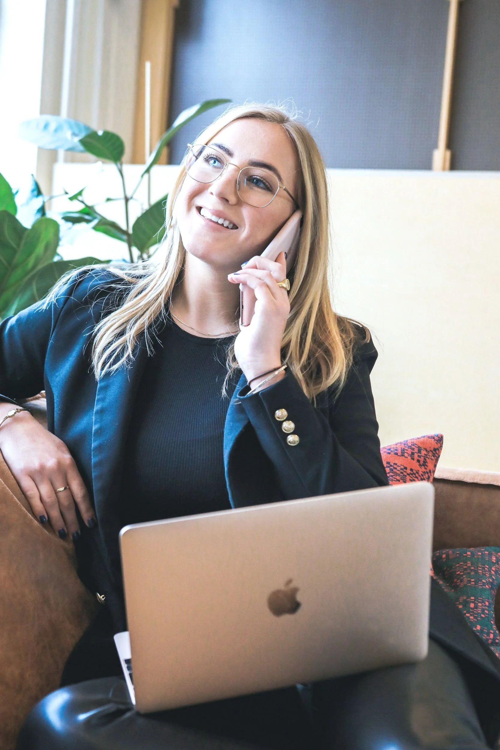 A white woman wearing a blazer is on a mobile phone call. She is smiling. An Apple laptop is on her lap.