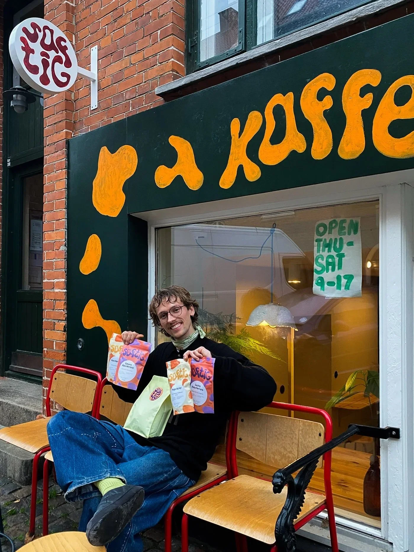 A young man with glasses and a black sweatshirt sits on the sidewalk outside a coffee shop, smiling and holding three bags of coffee beans.