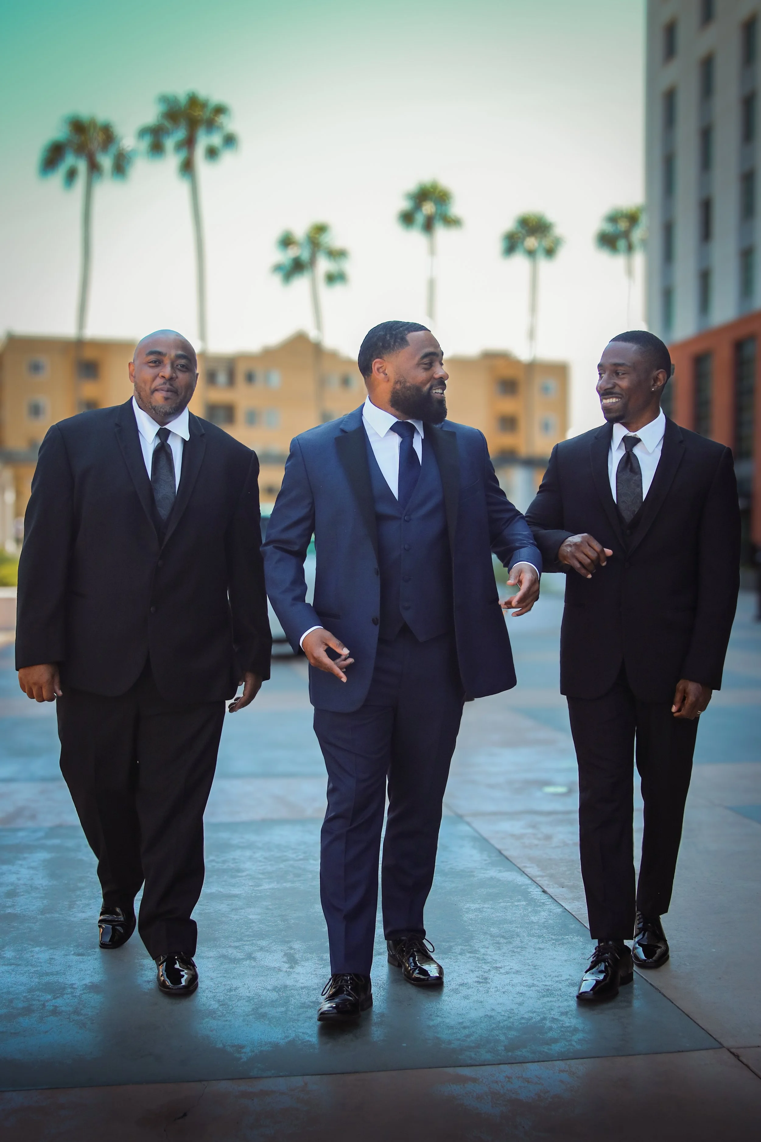 Three men in suits walking outdoors, smiling, with palm trees and buildings in the background.