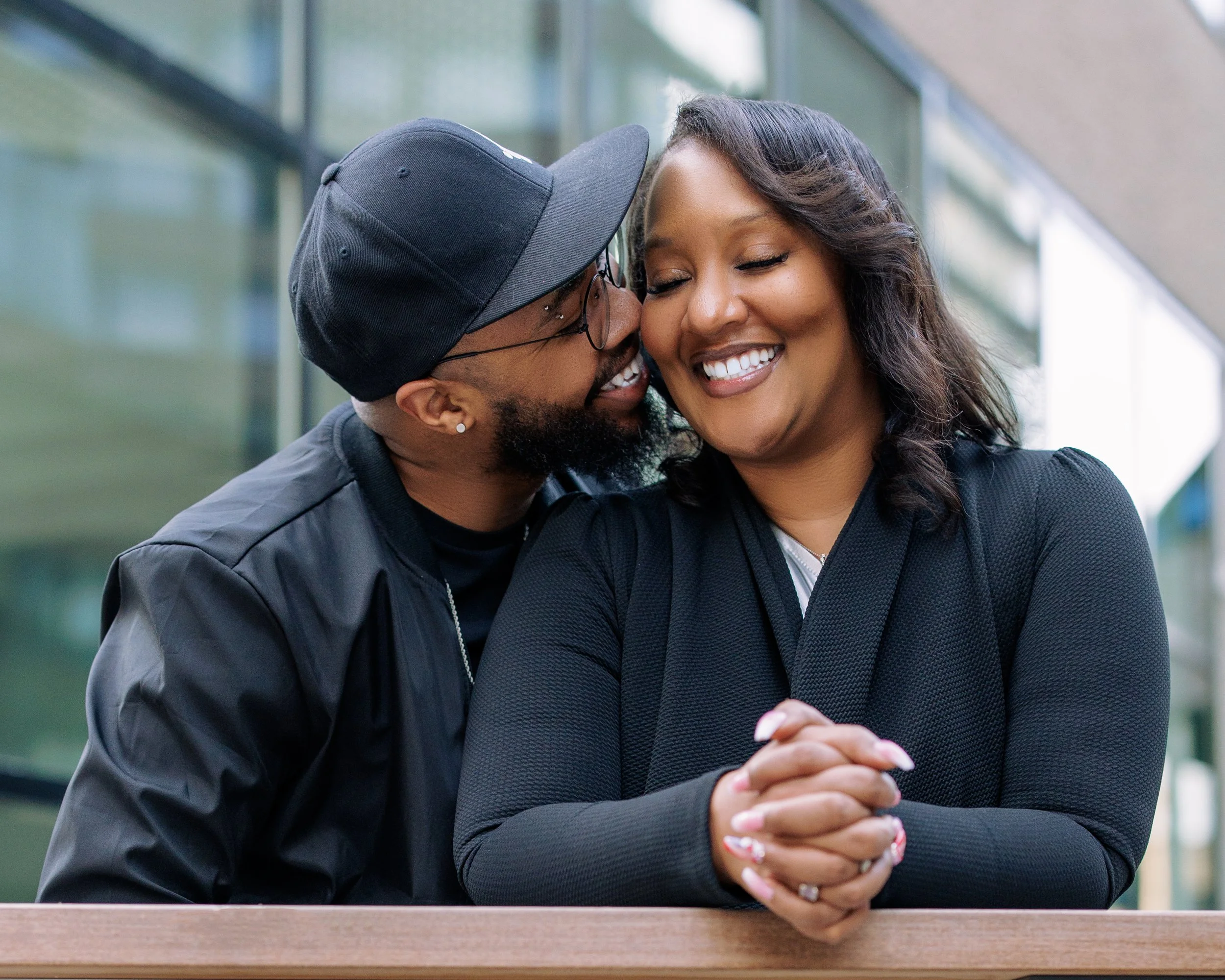 A smiling couple, a man and a woman, sharing a private moment outside a modern building. The man is leaning in close, whispering or speaking softly to the woman, who is smiling with her hands clasped. The man wears glasses, a black cap, and a black jacket. The woman has shoulder-length dark hair and is wearing a black top or blazer.