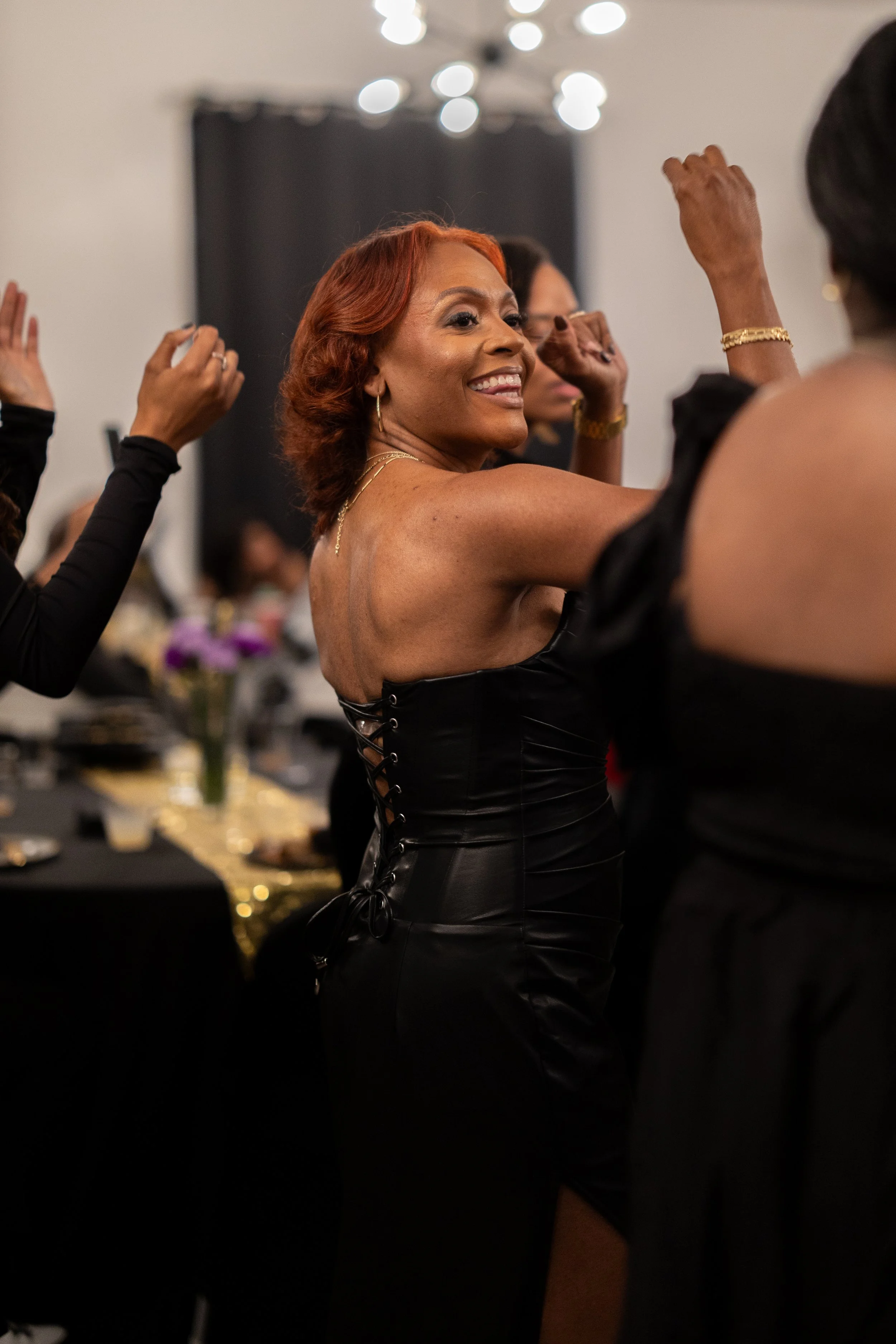 Woman with red hair dance at black-tie event, smiling and raising arms, surrounded by others in black attire, decorated table with flowers in background.