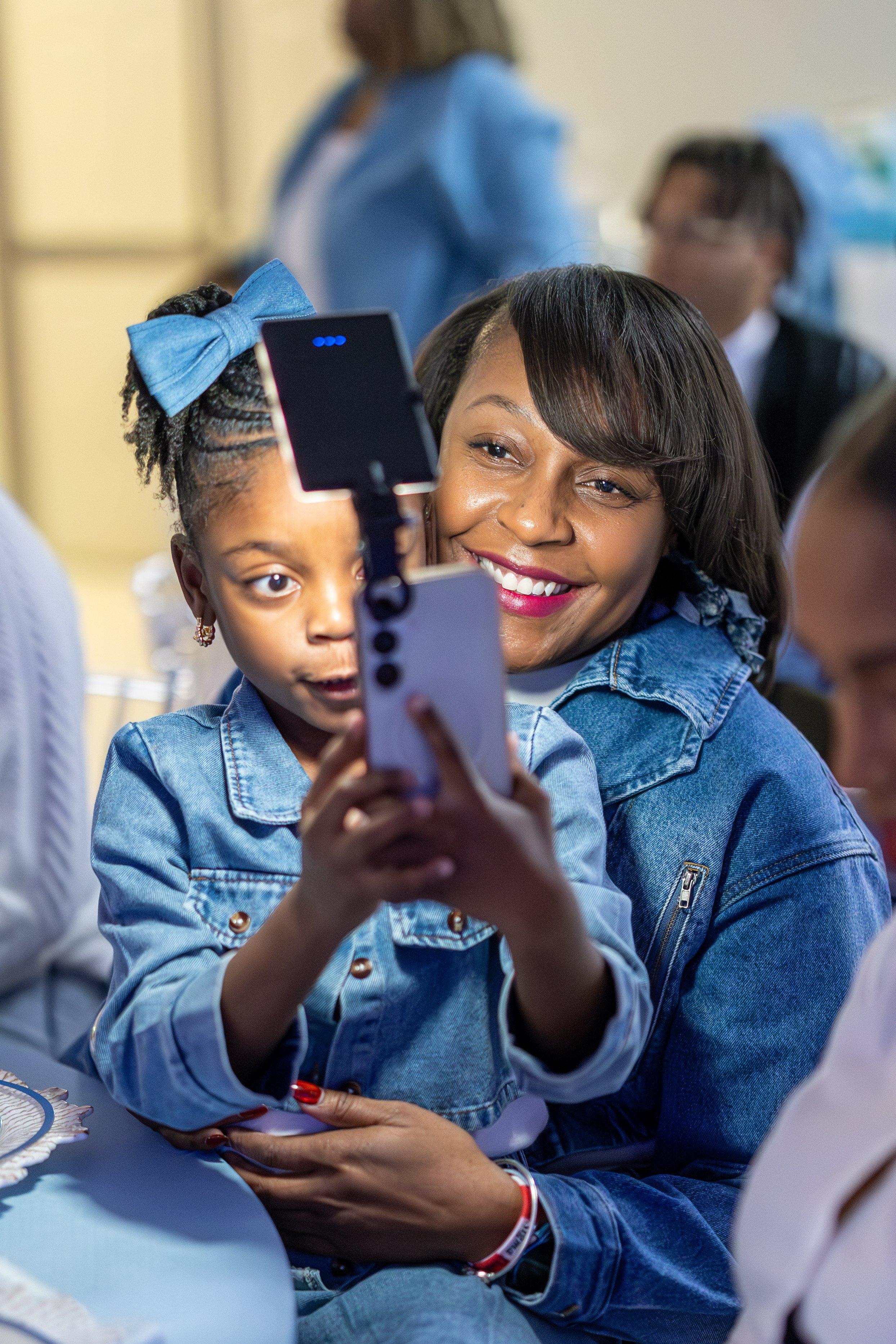 A woman and a young girl are taking a selfie together at an event. Both are wearing denim jackets, and the girl has a blue bow in her hair.