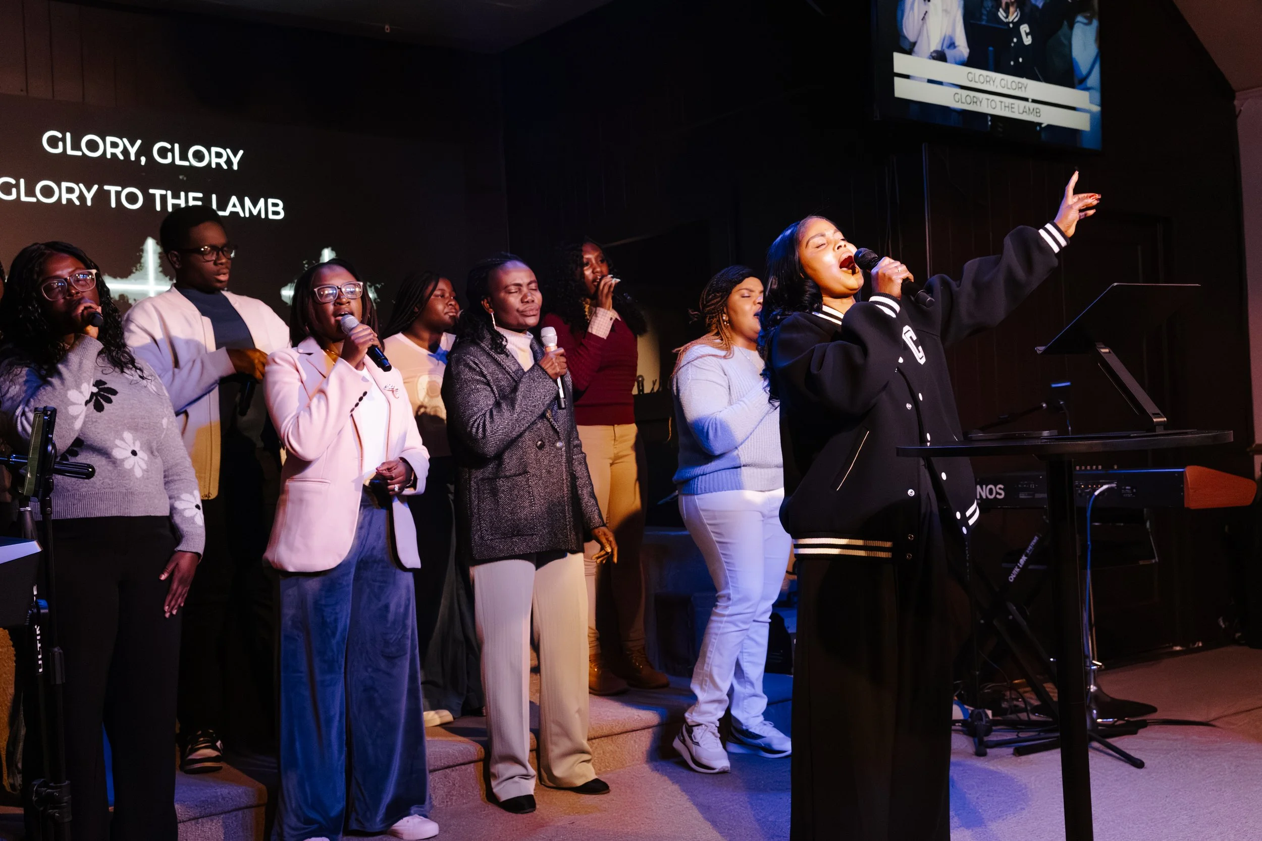 A group of diverse people singing on stage with a woman in front leading, lyrics 'Glory, Glory, Glory to the Lamb' projected behind them.