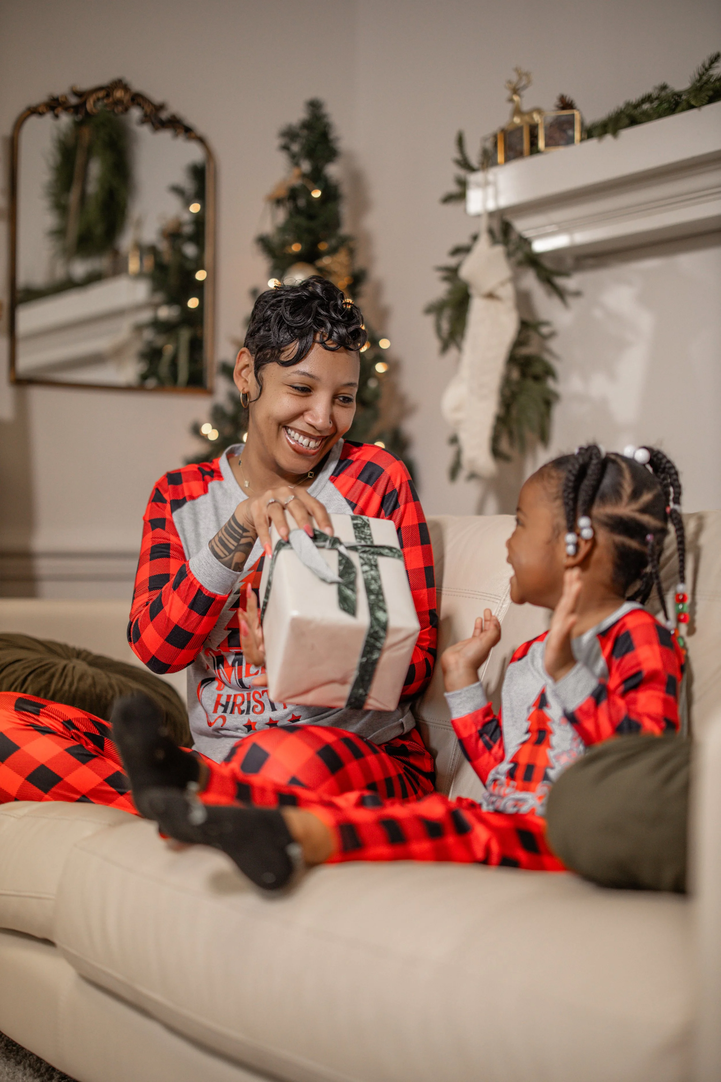 A woman and a young girl, both wearing matching Christmas pajamas, sit on a beige couch in a decorated living room. The woman is giving the girl a wrapped Christmas gift, and they are smiling at each other. The background features a Christmas tree, a