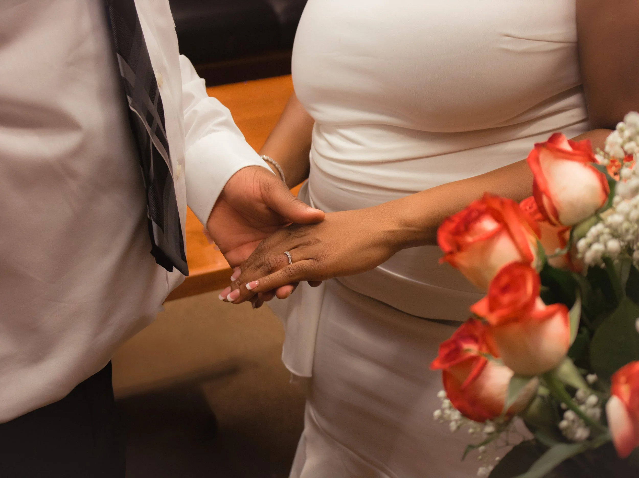 A couple holding hands during a wedding ceremony, with the bride holding a bouquet of pink and white roses.