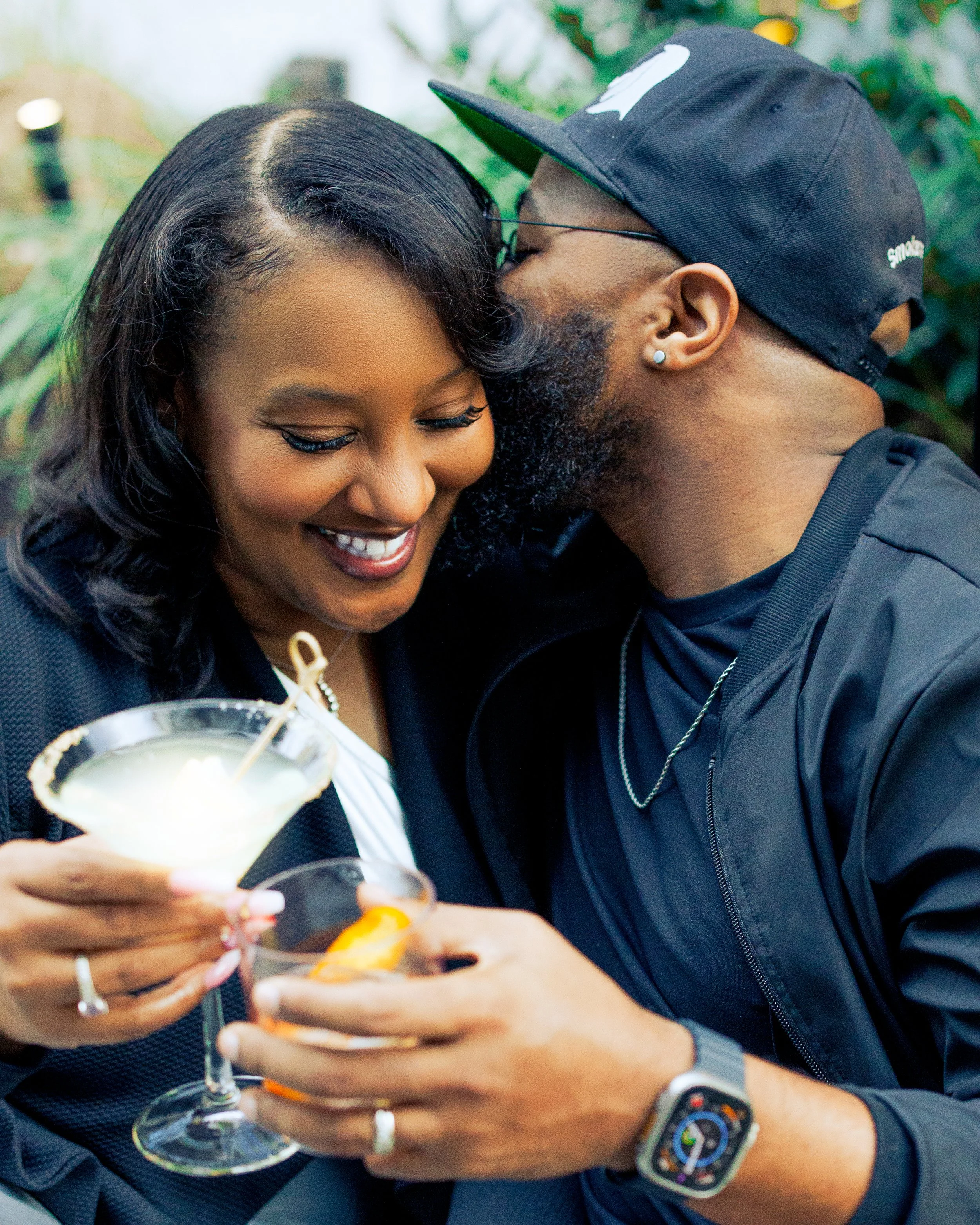 A smiling woman and a man share a close moment, holding cocktails, with the man whispering into the woman's ear, in a green, outdoor setting.