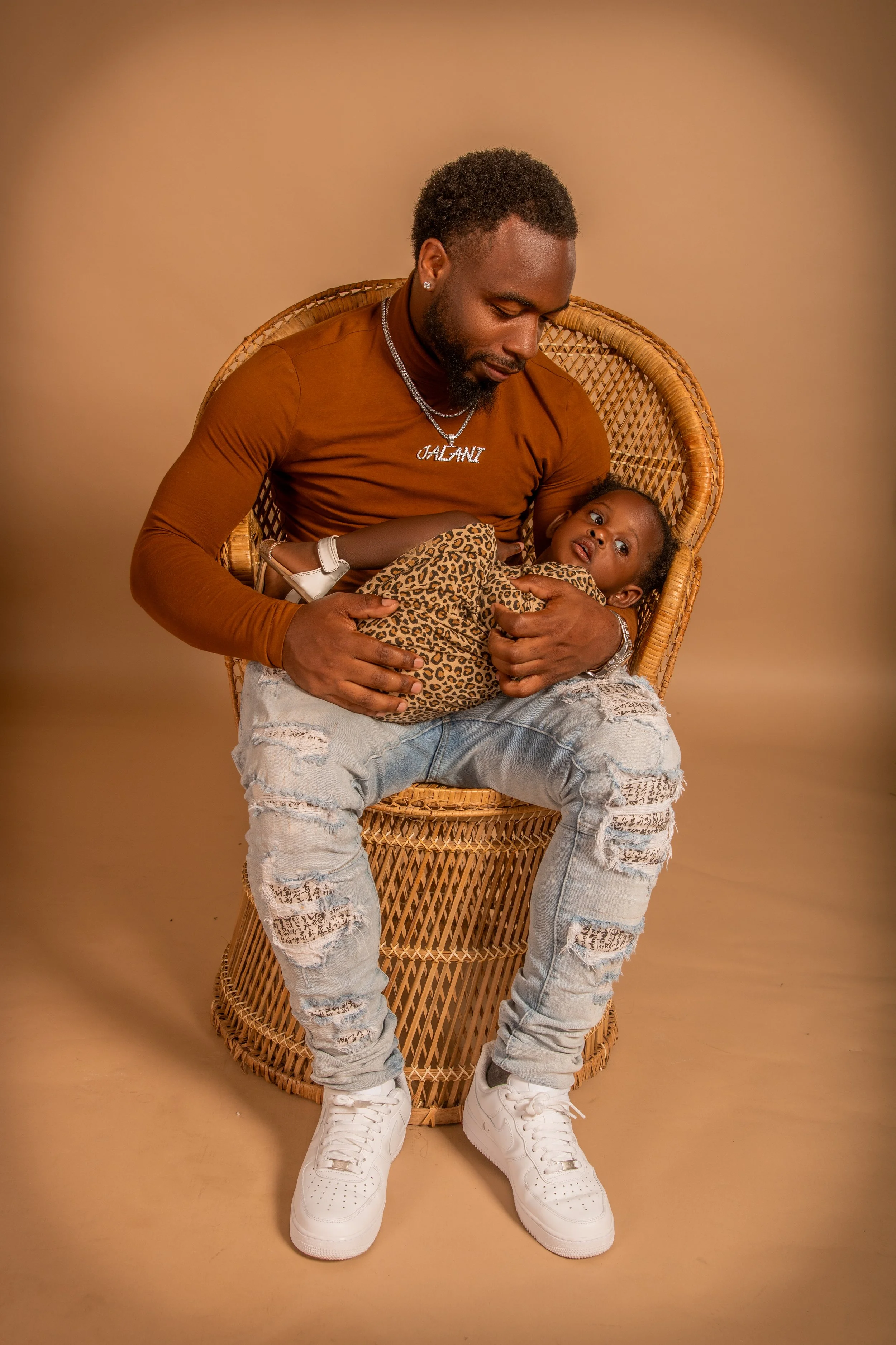 A man sitting on a wicker chair holding a young girl on his lap. The girl is wearing a leopard print outfit and looking up. The man is wearing a brown long sleeve shirt, distressed jeans, and white sneakers, with a necklace that says 'JALANI'.