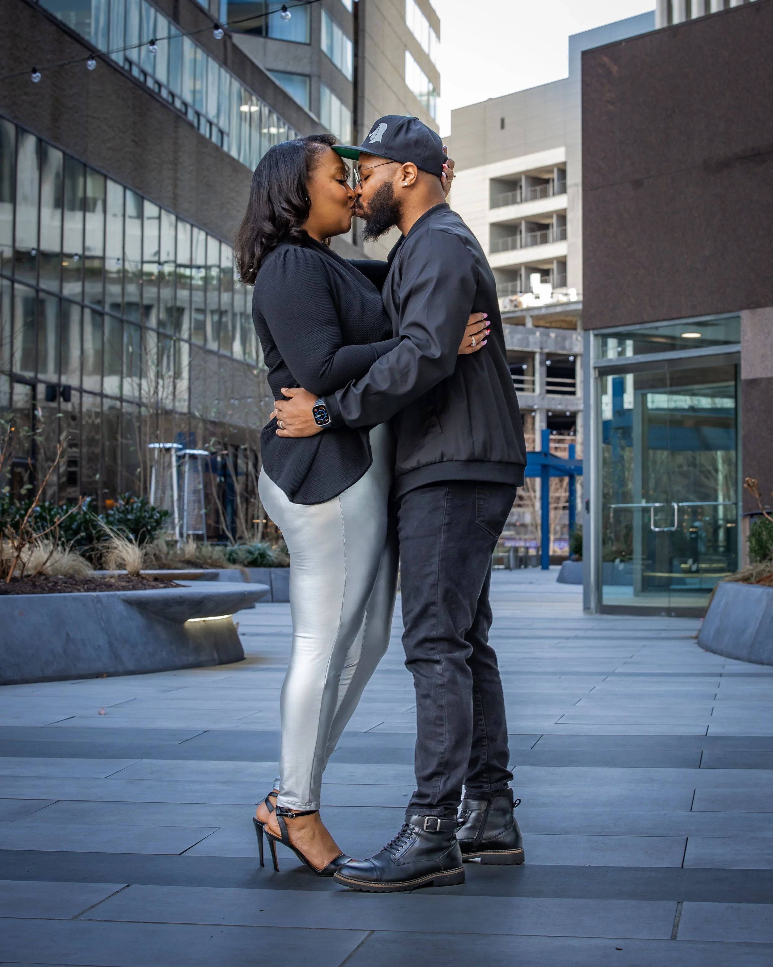 A couple is standing close, about to kiss, in an urban outdoor setting with modern buildings and planters.