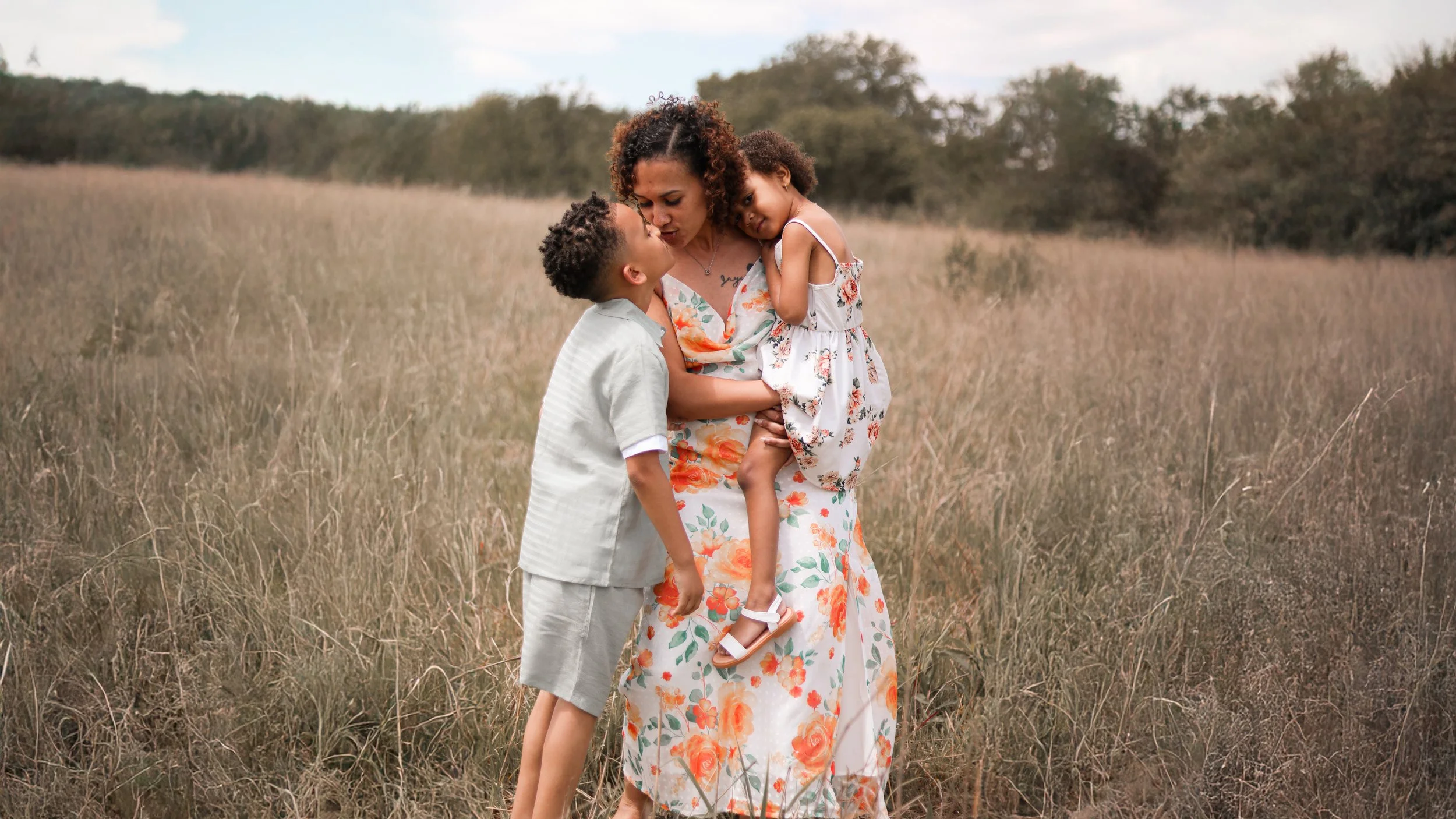 A woman holding two children in a grassy field during daytime, with trees in the background.