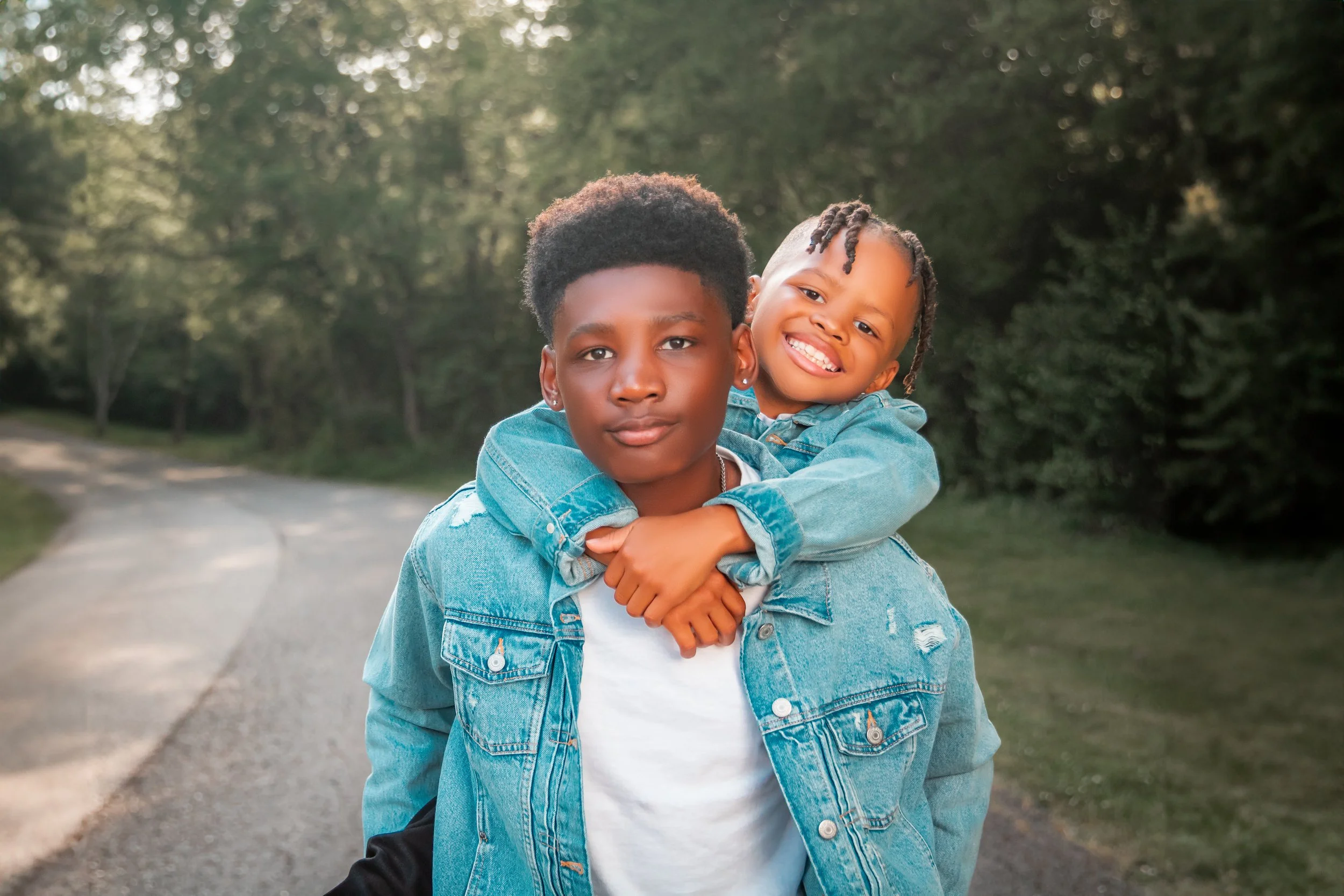 Two children, an older boy and a younger boy, are outdoors on a forest path during daylight. The little brother is on the big brother's back, hugging him around the neck, and both are smiling happily. They are both wearing denim jackets.