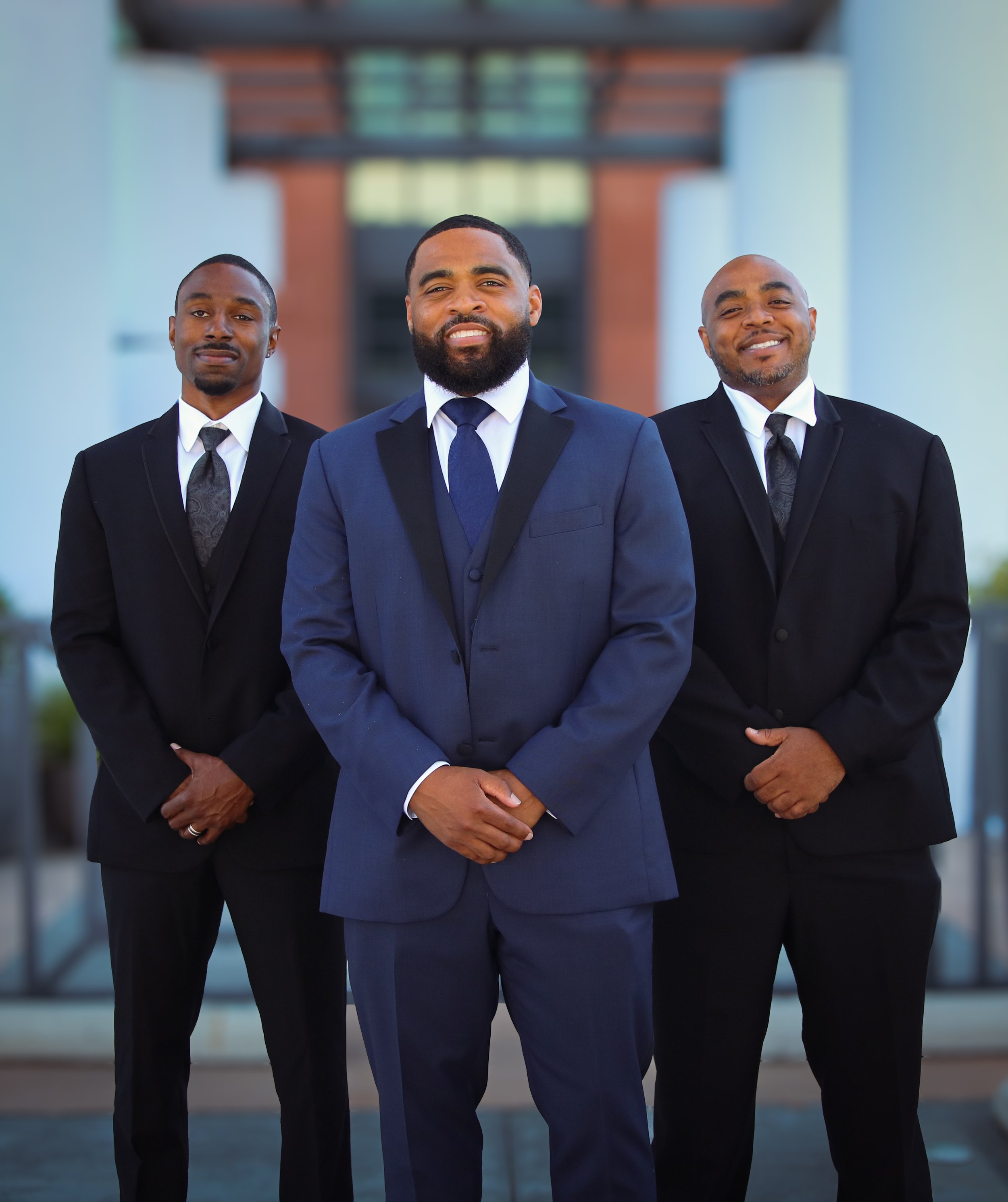 Three men in suits standing outdoors, smiling, with a building in the background.