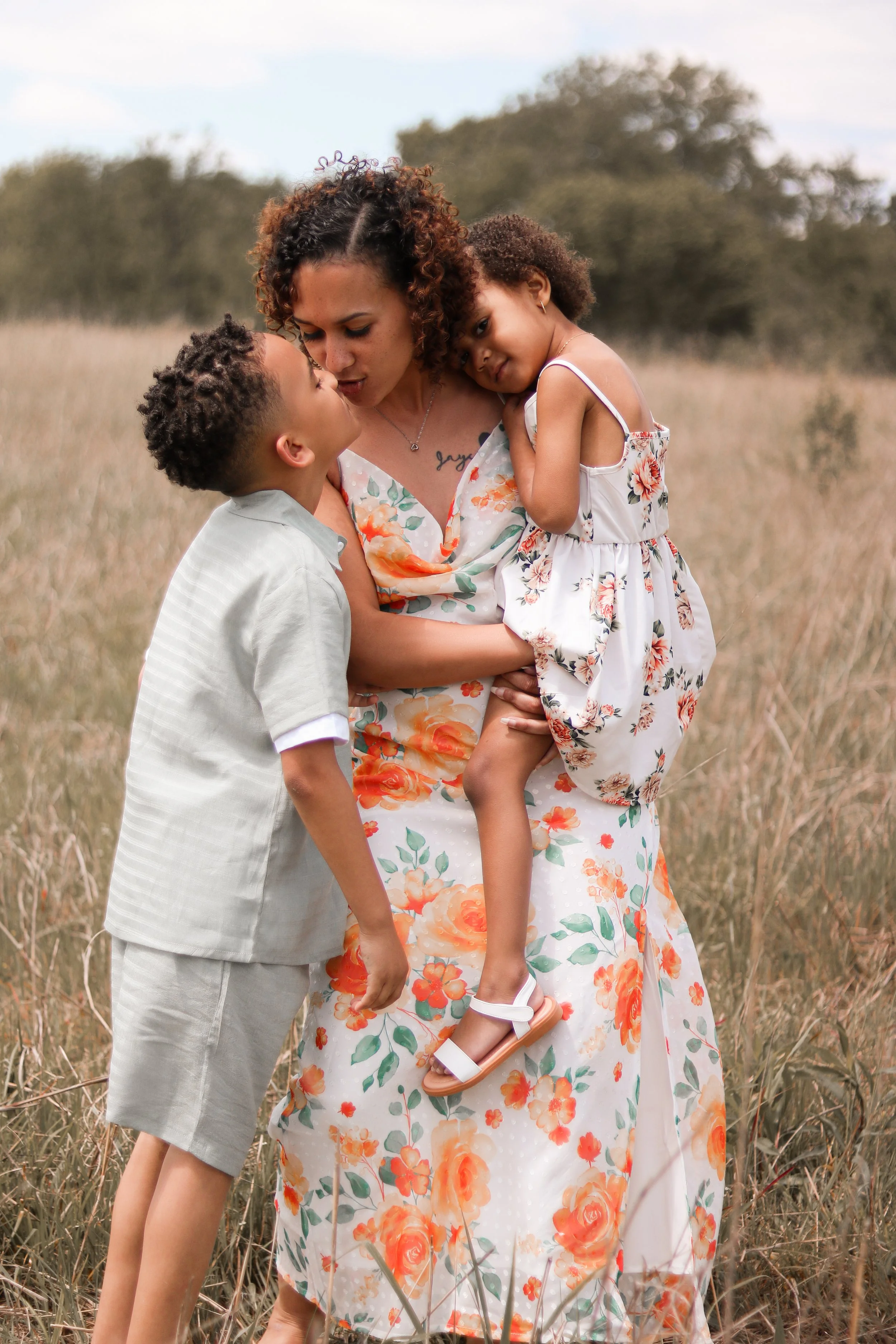 A woman holding a girl in a floral dress, a boy pressing his face close to her face, in an outdoor grassy field.