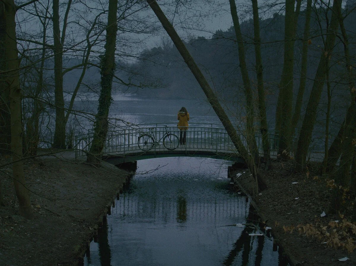 Still from A Letter to Home (2026), cinematography by Dorothea Pilz: a figure in a red coat on a small bridge in a misty winter park at dusk