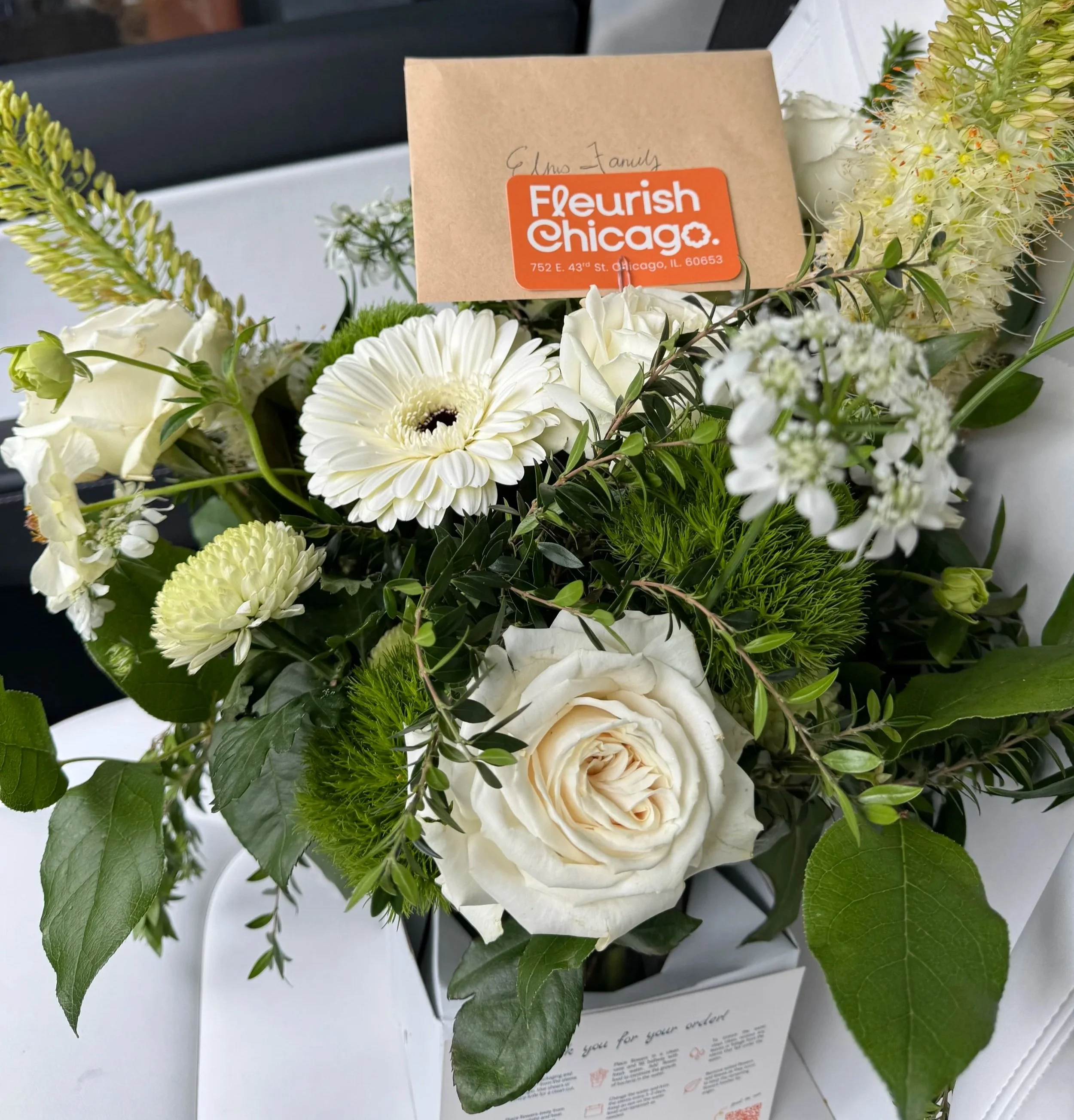 A bouquet of white and green flowers with a card that reads 'Fleurish Chicago, 752 E. 43rd St., Chicago, IL 60653' and a handwritten note that says 'Elmo Family'.