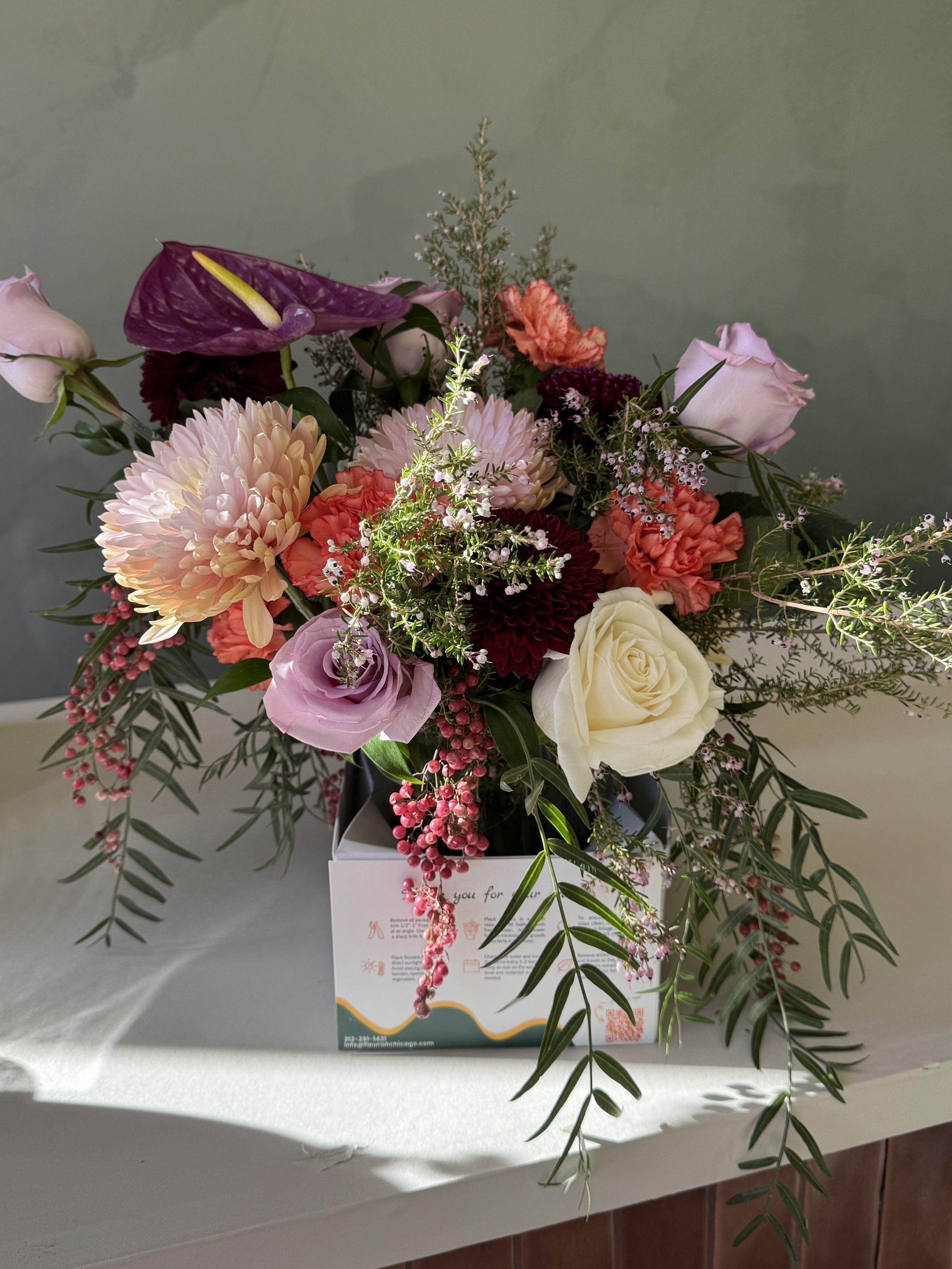 A floral arrangement in a white display box on a white surface, featuring pink, white, purple, and red flowers with greenery and small white accents.