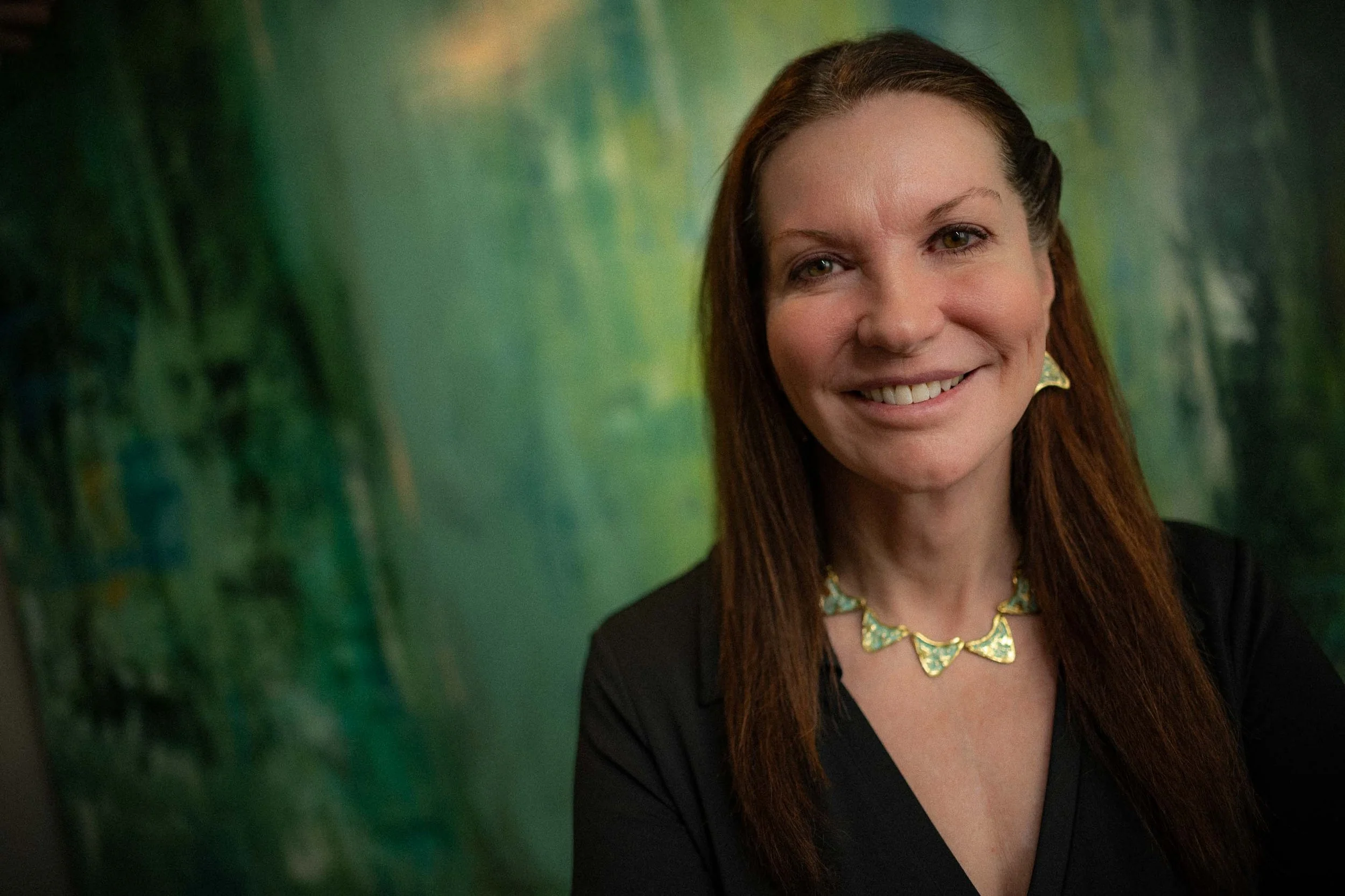 A woman with red hair smiling, wearing a black top and a gold and green necklace. The background appears to be an abstract green painting or mural.