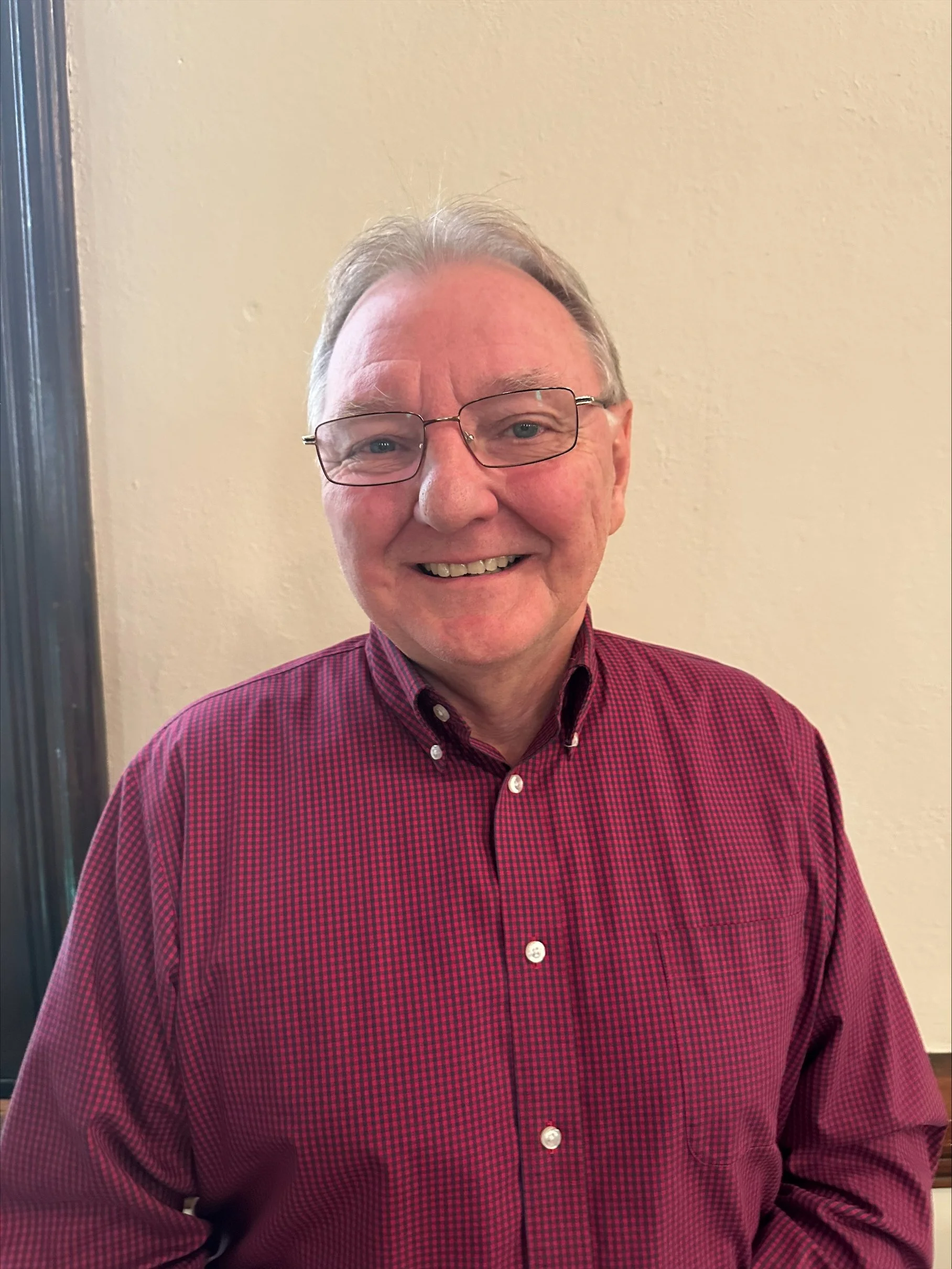 A smiling middle-aged man wearing glasses and a red checkered shirt, standing against a beige wall.