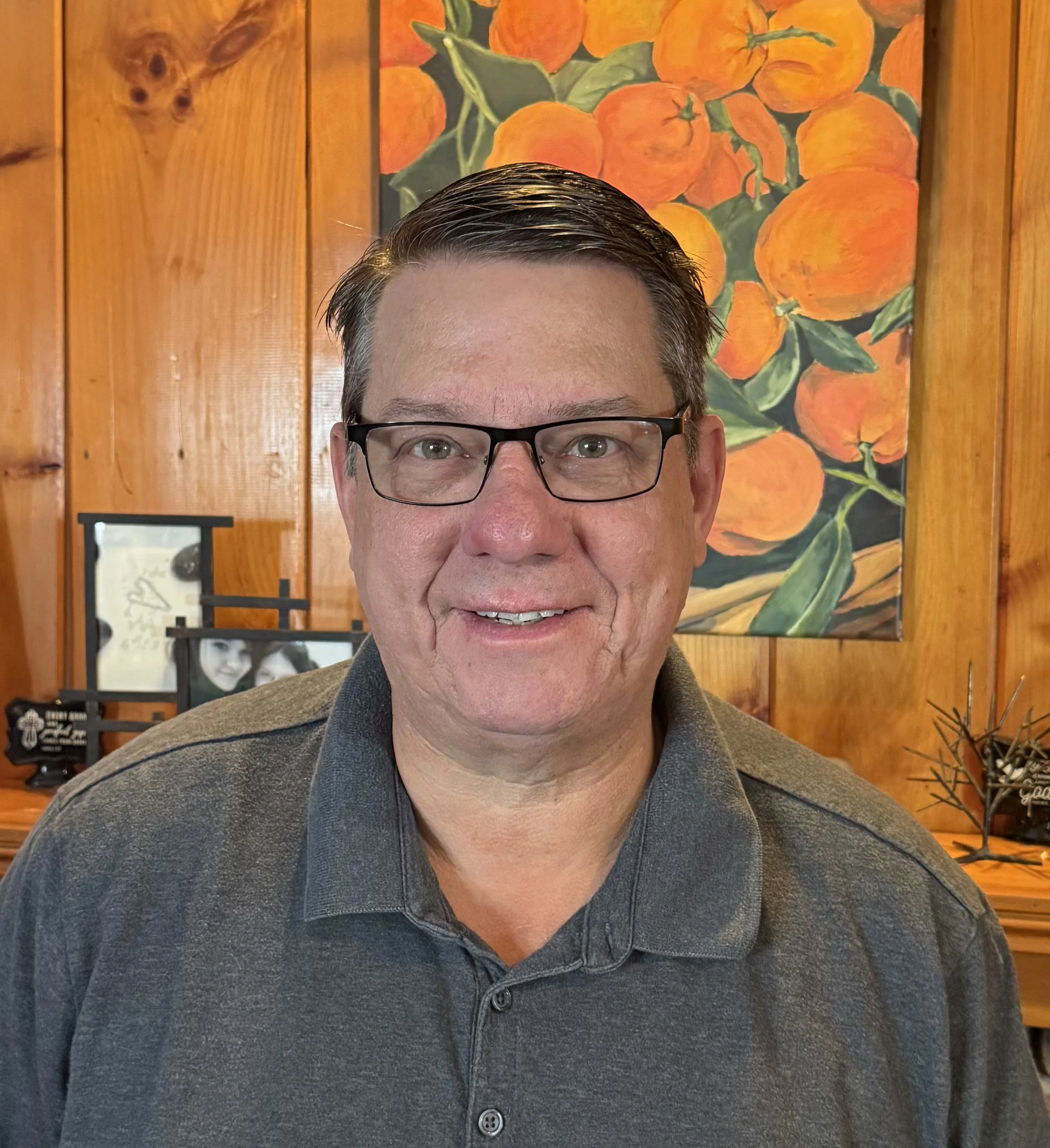 A middle-aged man with short dark hair, glasses, and a gray polo shirt, smiling indoors in front of a wooden wall with a painting of oranges and green leaves and a small photo frame in the background.