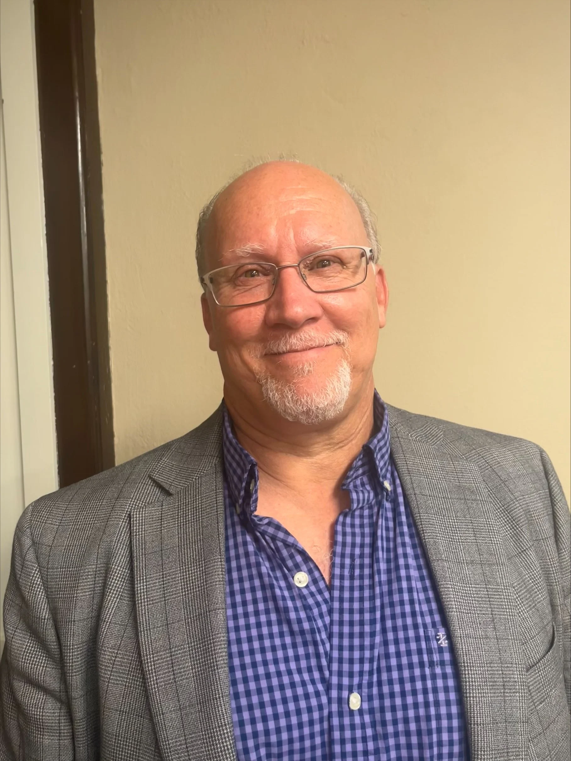 A middle-aged man with glasses, a goatee, and a bald head smiling, wearing a checkered blue shirt and a gray blazer, standing against a beige wall.
