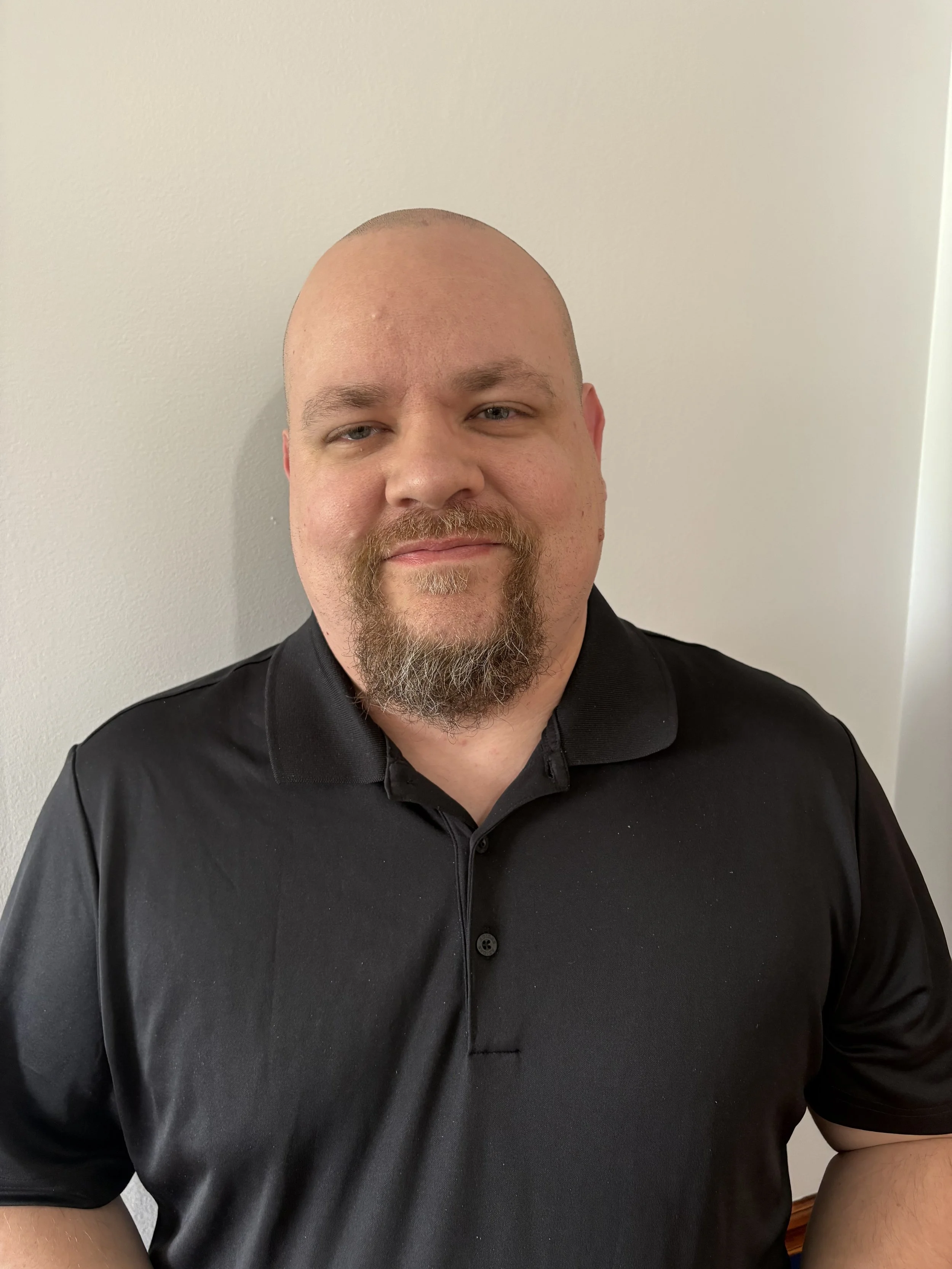 A man with a shaved head and beard smiling, wearing a black polo shirt, standing against a plain wall.