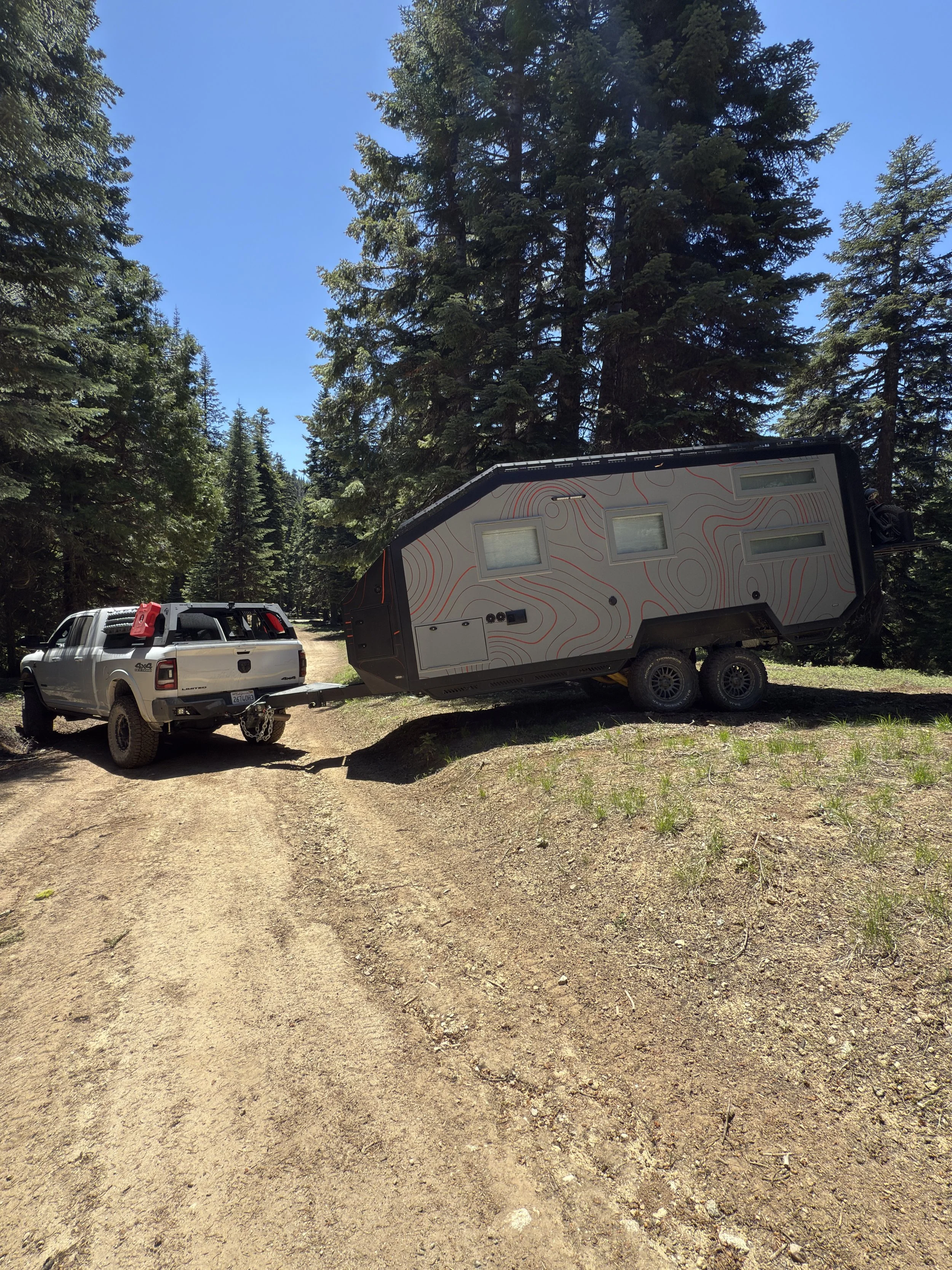 Interior of a tiny home kitchen with a black countertop, stainless steel sink, and wooden cabinets above and below. A window shows trees outside. Mounted on the wall are a TV, a tablet, and a monitor arm, with a small potted plant on the counter.