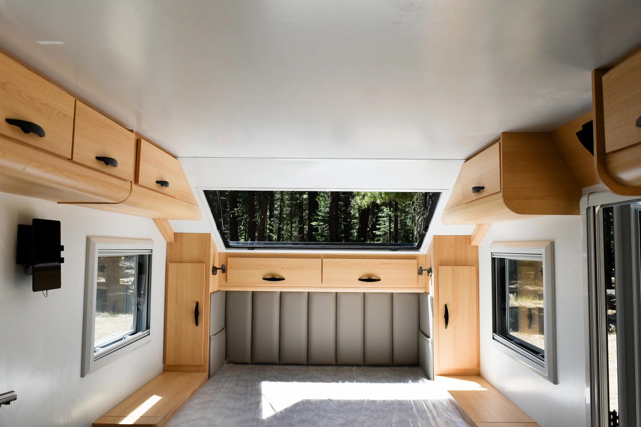 Interior of a camper with a bed, wooden cabinets, windows, and a large skylight showing trees outside.