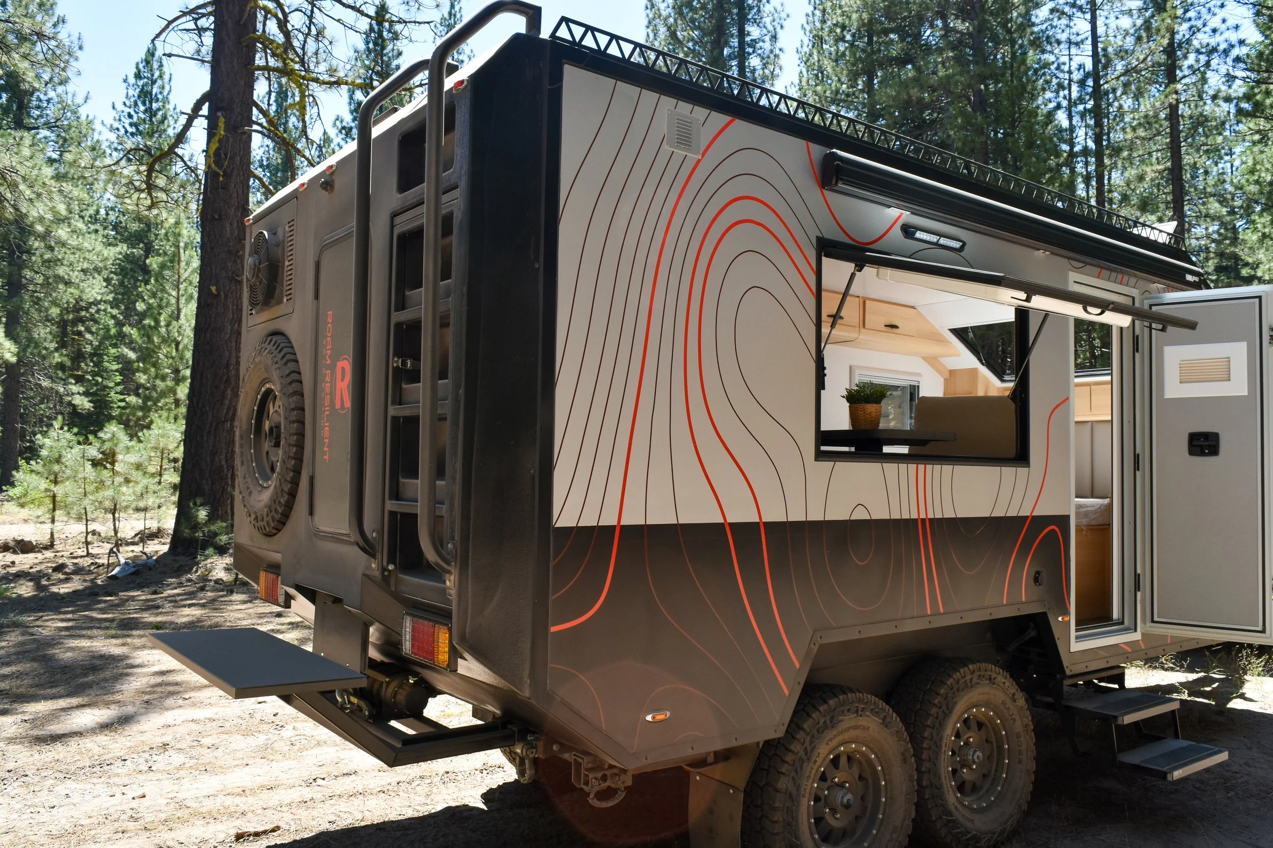 A modern off-road camper trailer parked in a wooded area with trees in the background, featuring a window, stairs, and outdoor amenities.