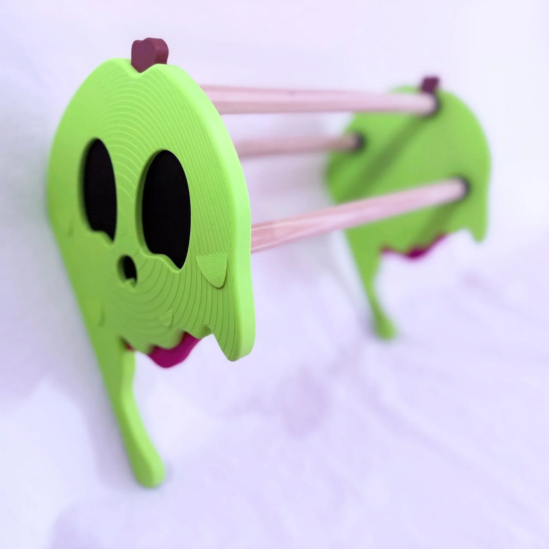 Two green wall hooks shaped like cartoon skulls with black eye sockets, mounted on a white wall, holding pink plastic sticks.