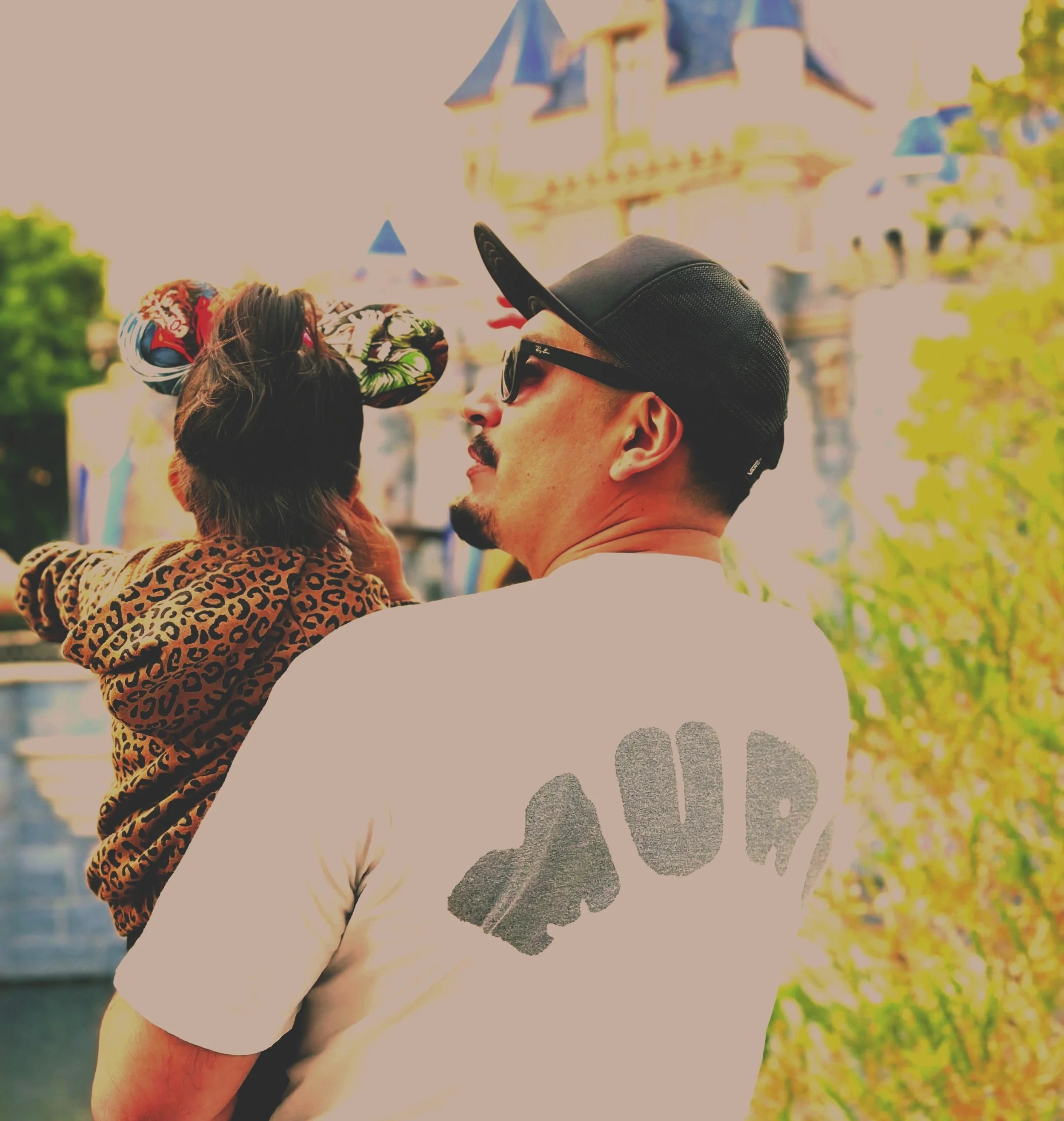 A man wearing sunglasses, a cap, and a white T-shirt holding a child with a leopard print hoodie, both looking at a colorful sculpture or statue in an outdoor park setting.