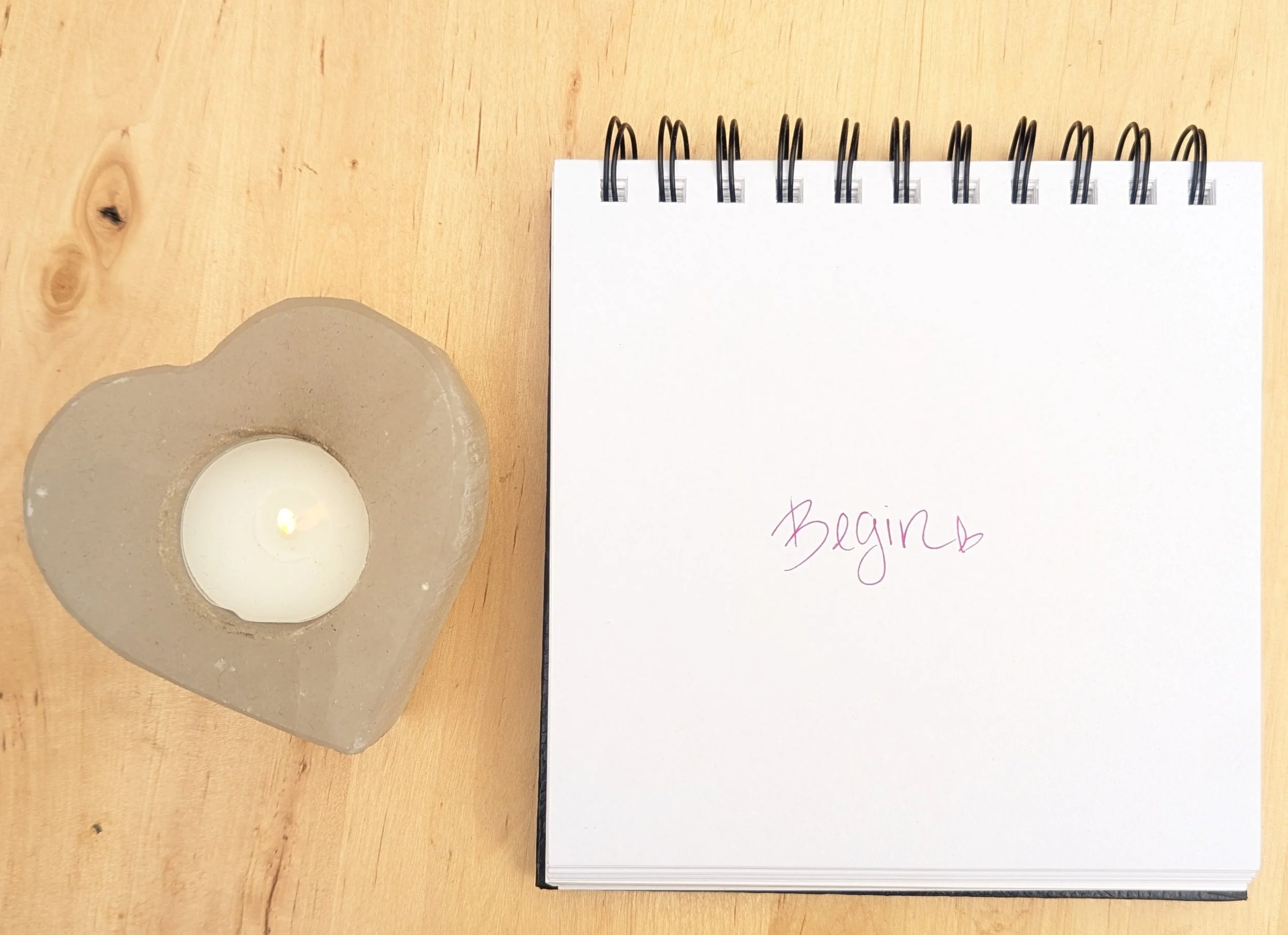 A spiral-bound journal with the handwritten word “Begin” and a small heart beside a lit candle in a heart-shaped holder on a wooden table.