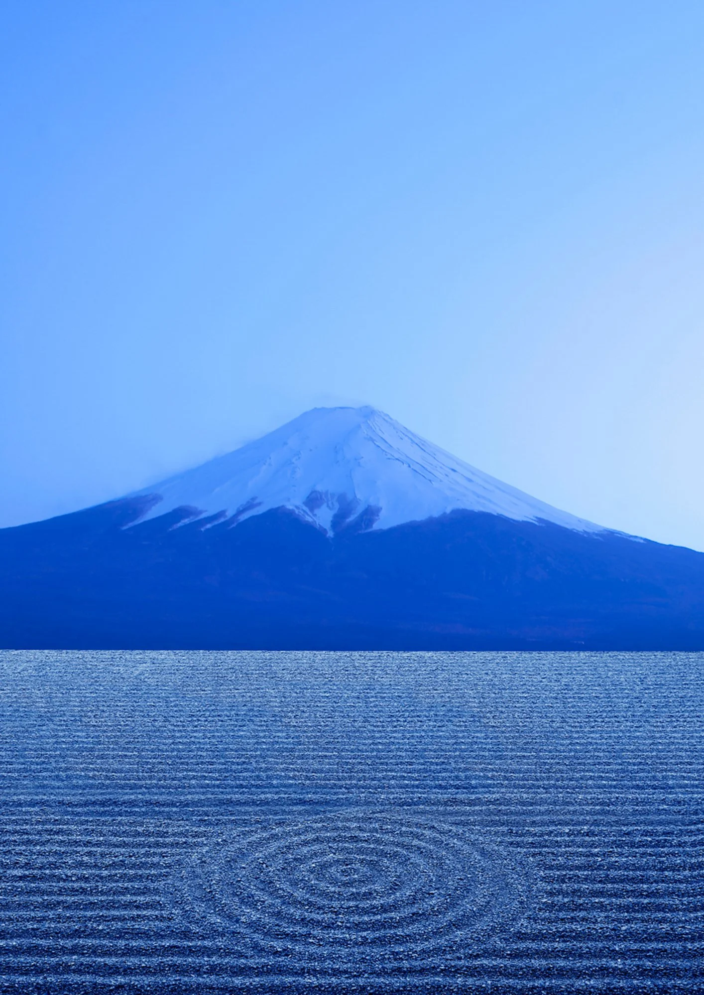 Blue image of a snow-capped mountain - Mount Fuji - reflected in a body of water with ripples making a spiral, under a clear blue sky.