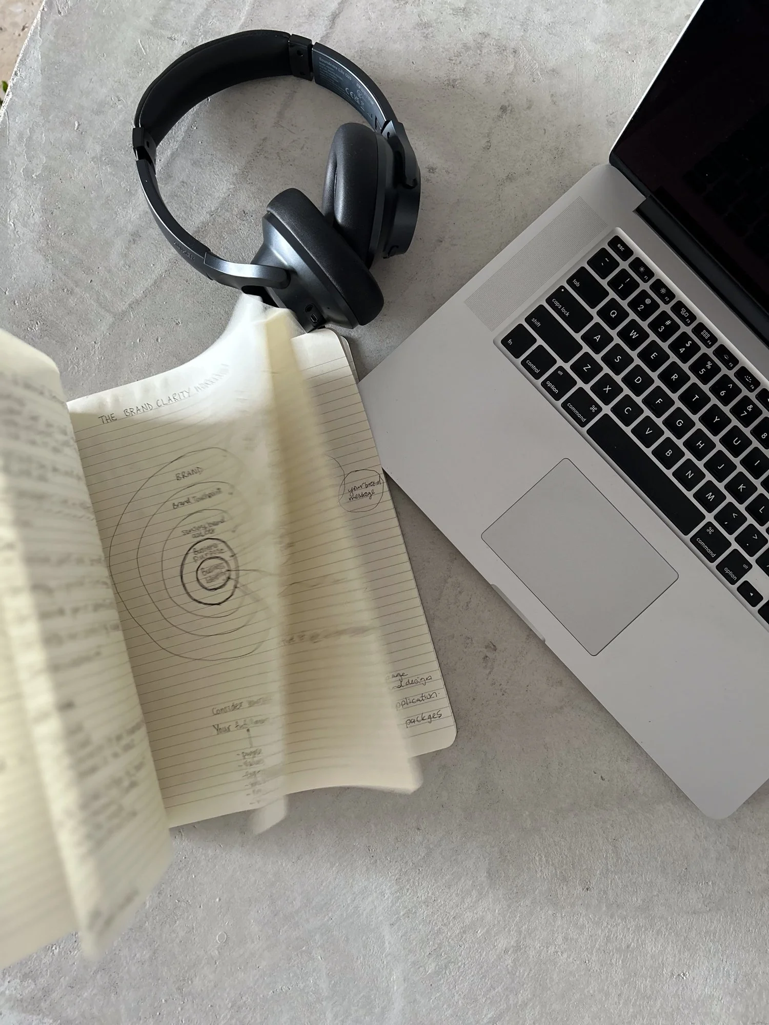 A workspace with a MacBook laptop, a notebook with handwritten notes and diagrams, a pair of black headphones, all placed on a light gray concrete surface.