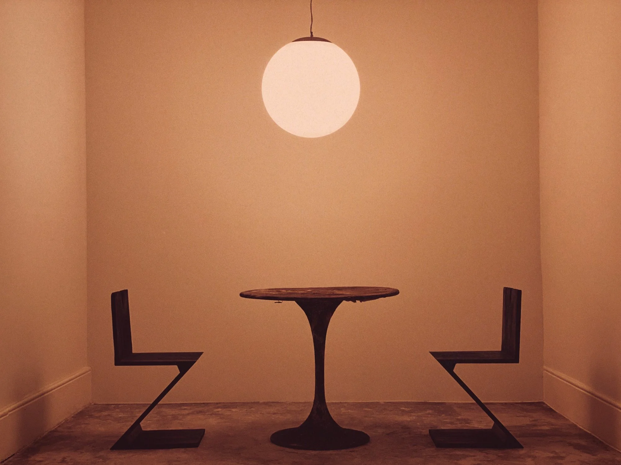 Orange image of a simple room, blank walls, dim lighting, a round hanging ceiling light, a small wooden table with a curved pedestal base, and two black metal chairs with angled legs and wooden seats.