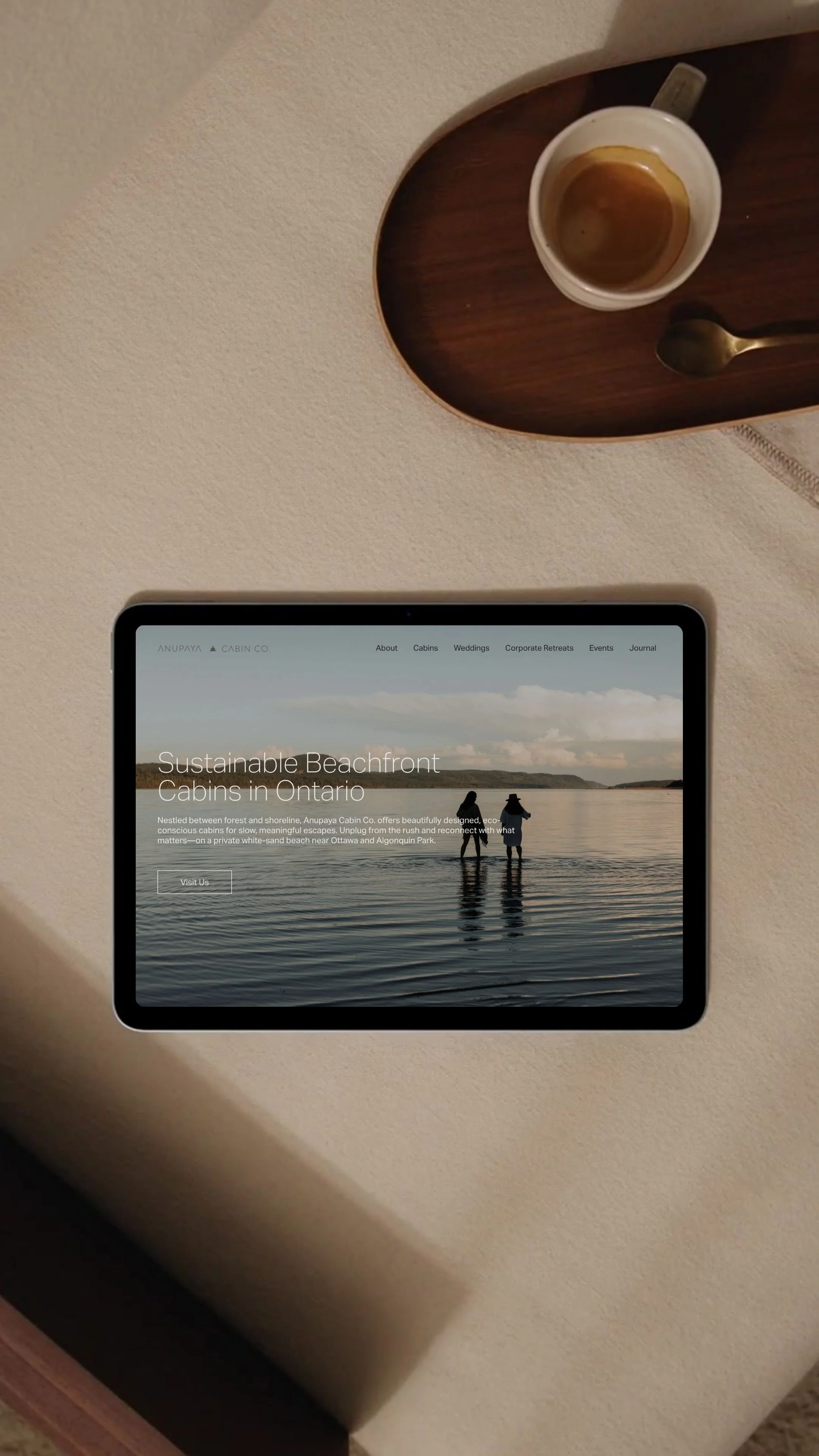 Tablet displaying a website for Anupaaya Cabin Co. with an image of two people walking in water on the beach. Beside the tablet is a cup of coffee on a dark wood tray.
