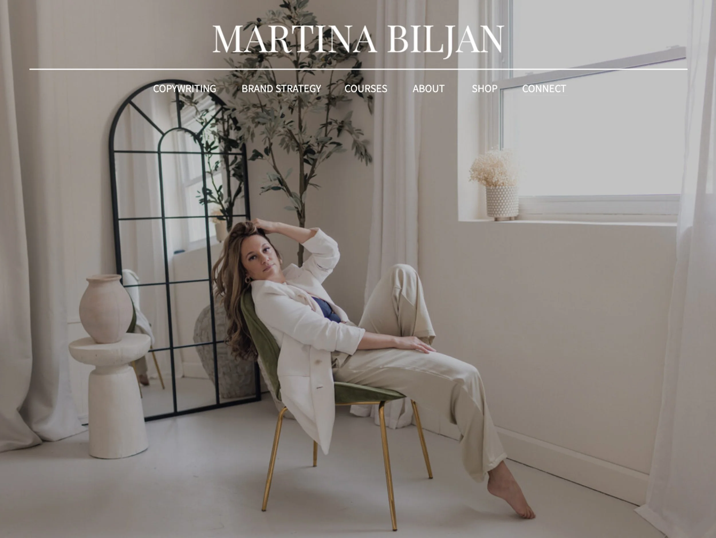 Website home page section with large 'Martina Biljan' wordmark logo is centred on top of the page with navigation links below on top of a background photo of Martina leaning back in a chair in a room with a window, large mirror and ceramic vase.