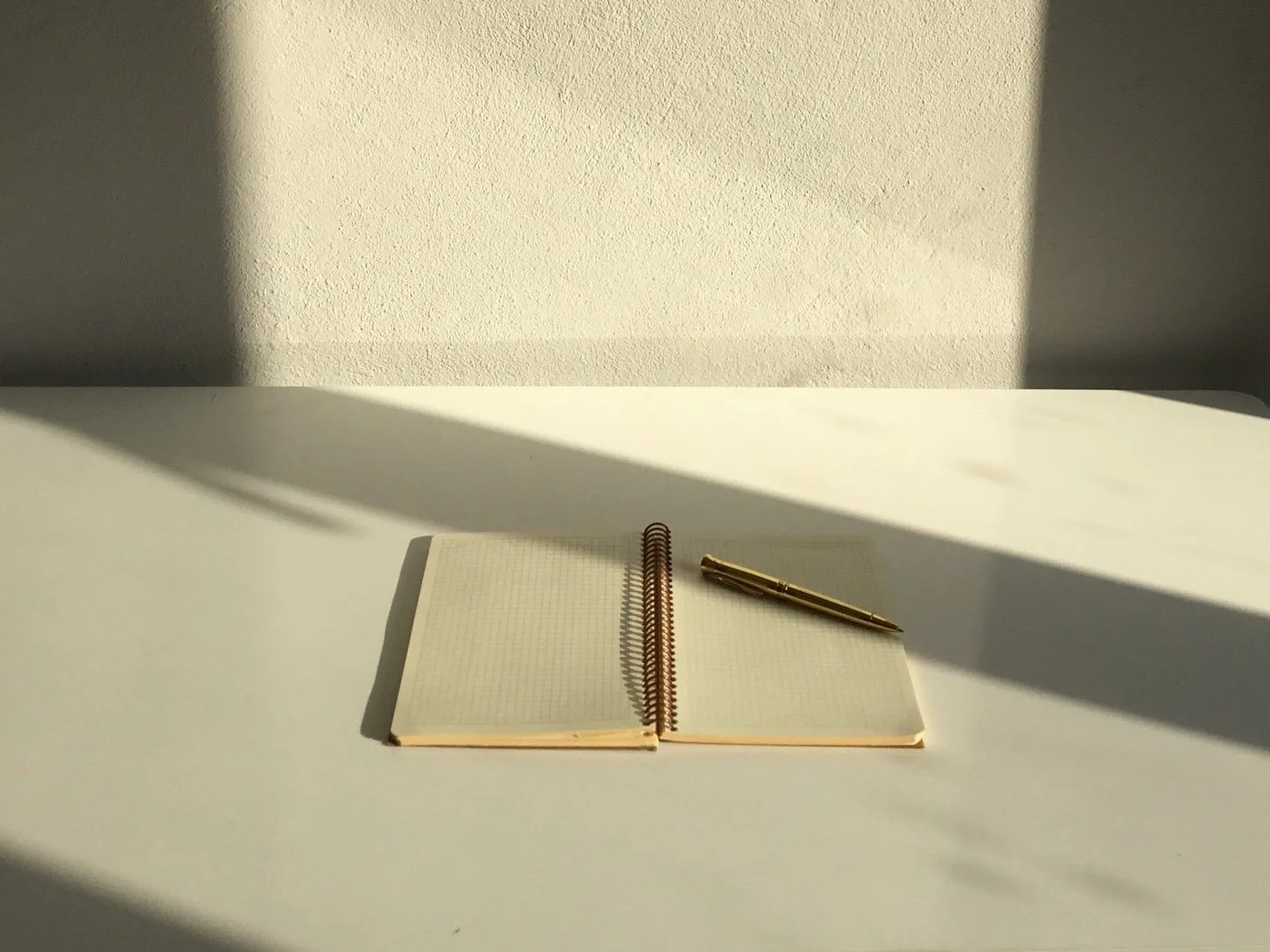 An open spiral-bound notebook with a gold pen resting on it, placed on a white surface, with sunlight creating shadows on the surface and the wall behind.