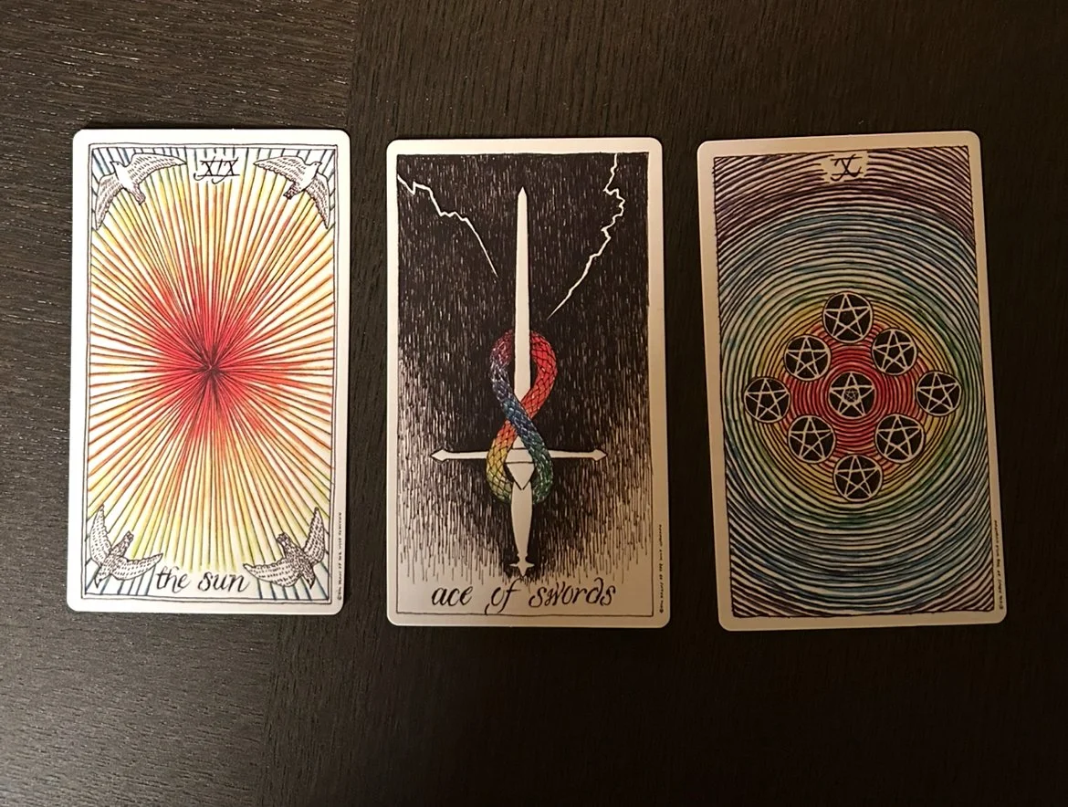 Three tarot cards: The Sun, Ace of Swords, and a card with concentric rainbow circles and seven pentacles.