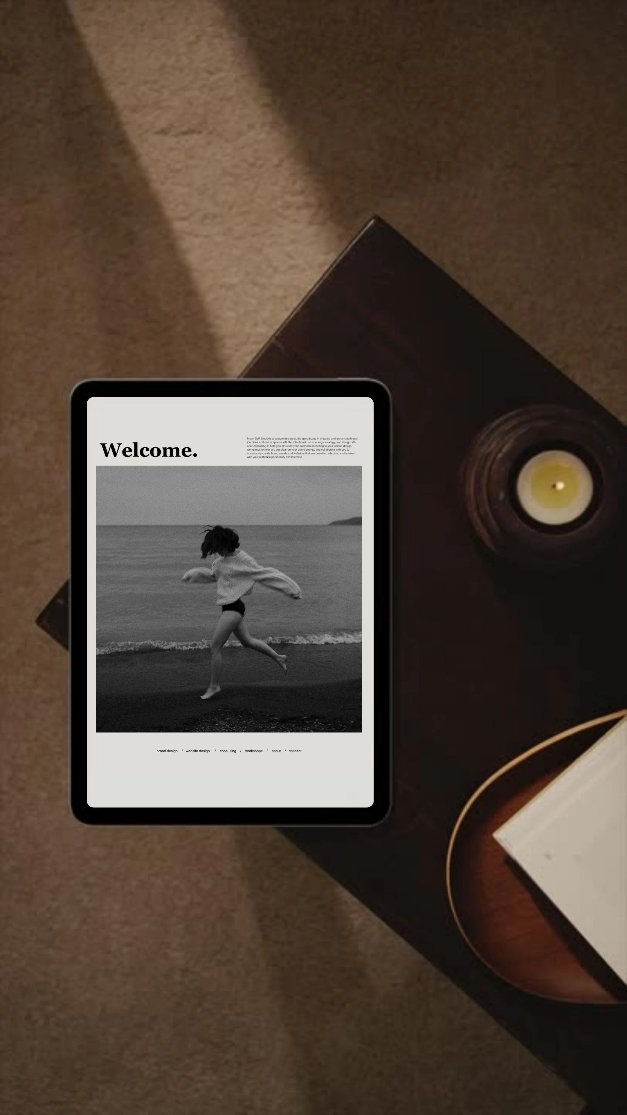 A tablet displaying an editorial website home page with the word welcome in larger text above a black and white photo of a girl jumping on the beach. The tablet is on a dark wooden table with a tealight candle beside.