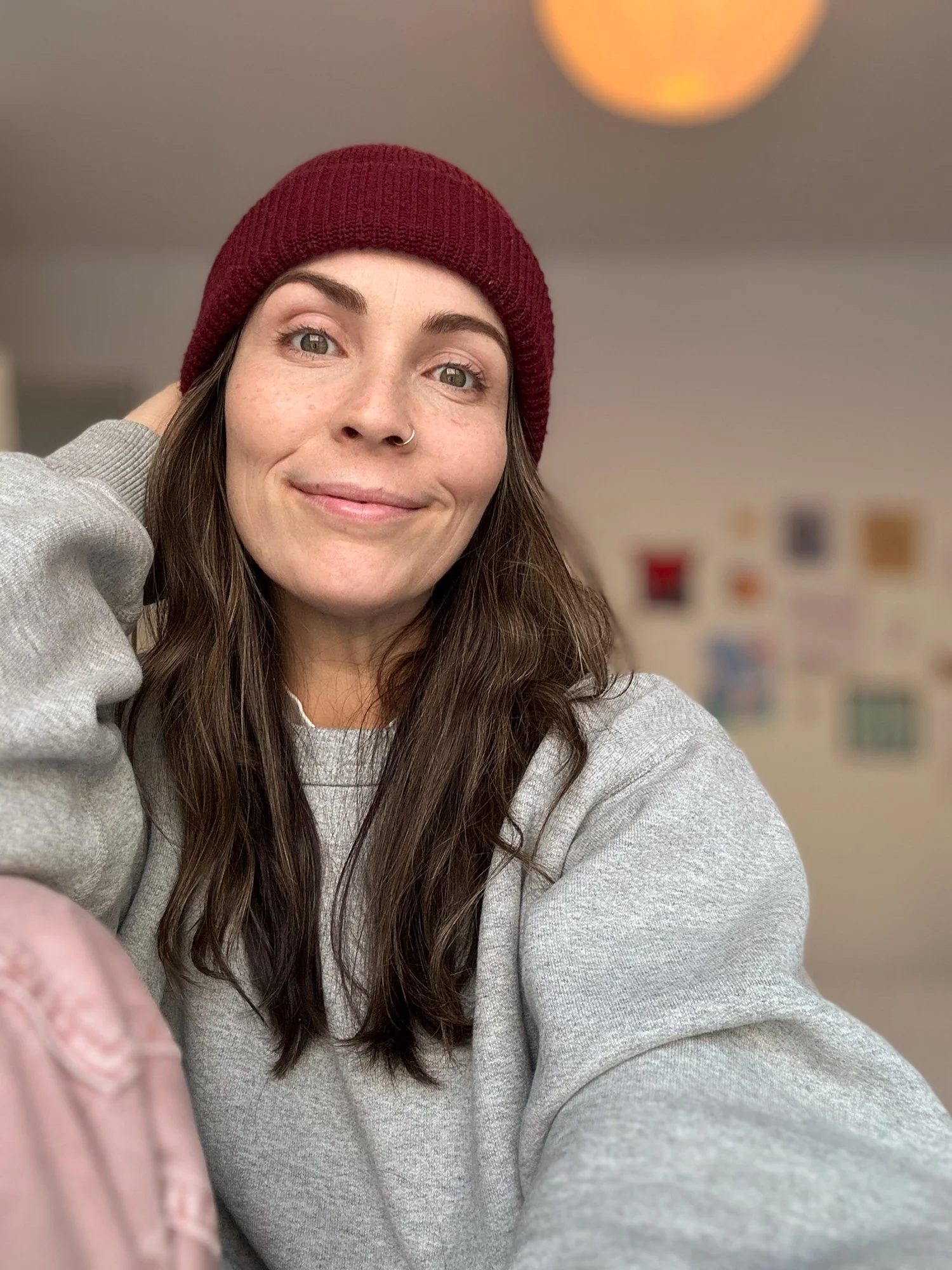 Steph Thornton with long brown hair, wearing a gray sweatshirt and a burgundy beanie, smiling at the camera in a room with a blurred background of colorful photos or posters.