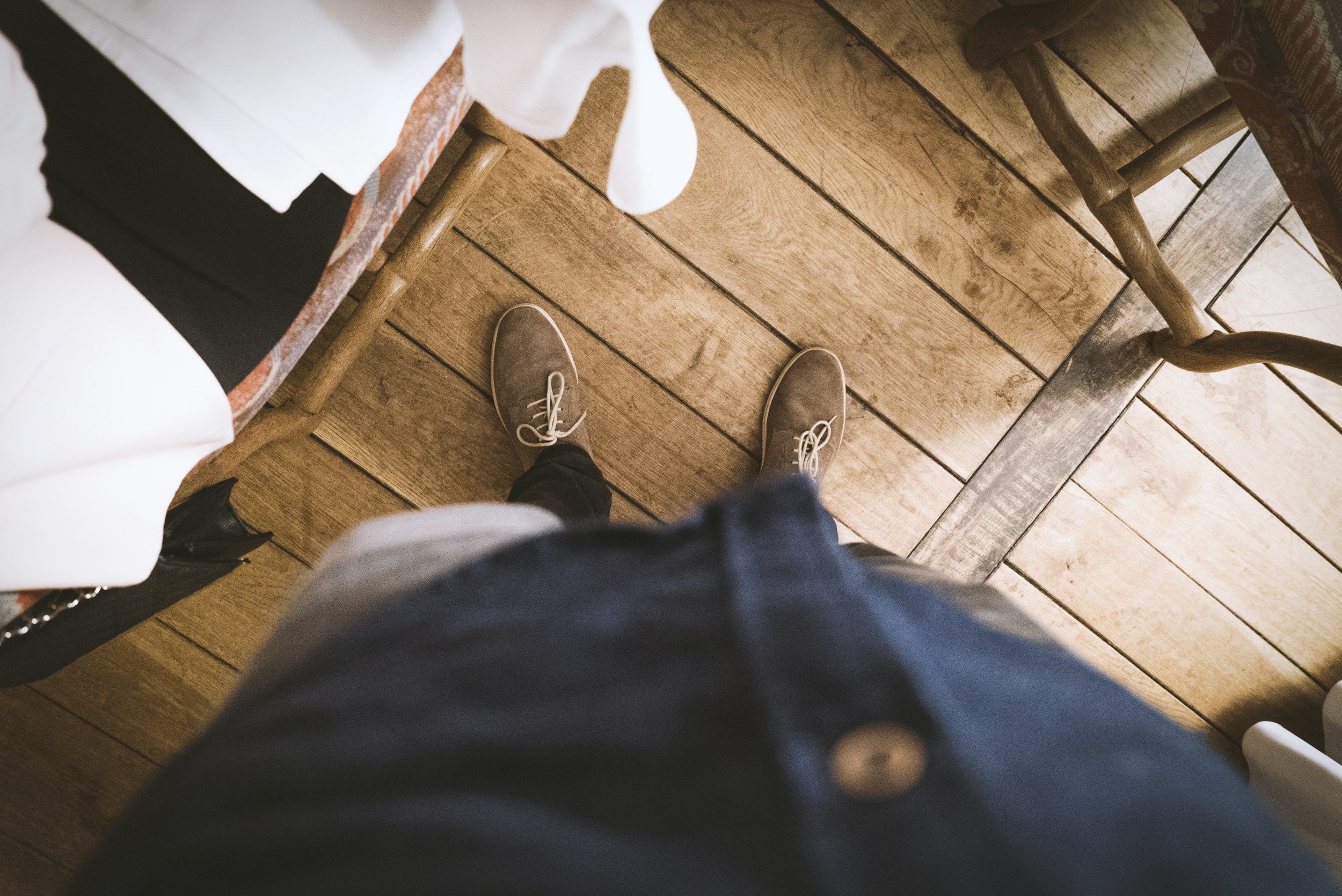 Overhead view of a person standing on a wooden floor wearing beige shoes and dark pants, with part of a white tablecloth and chairs visible.