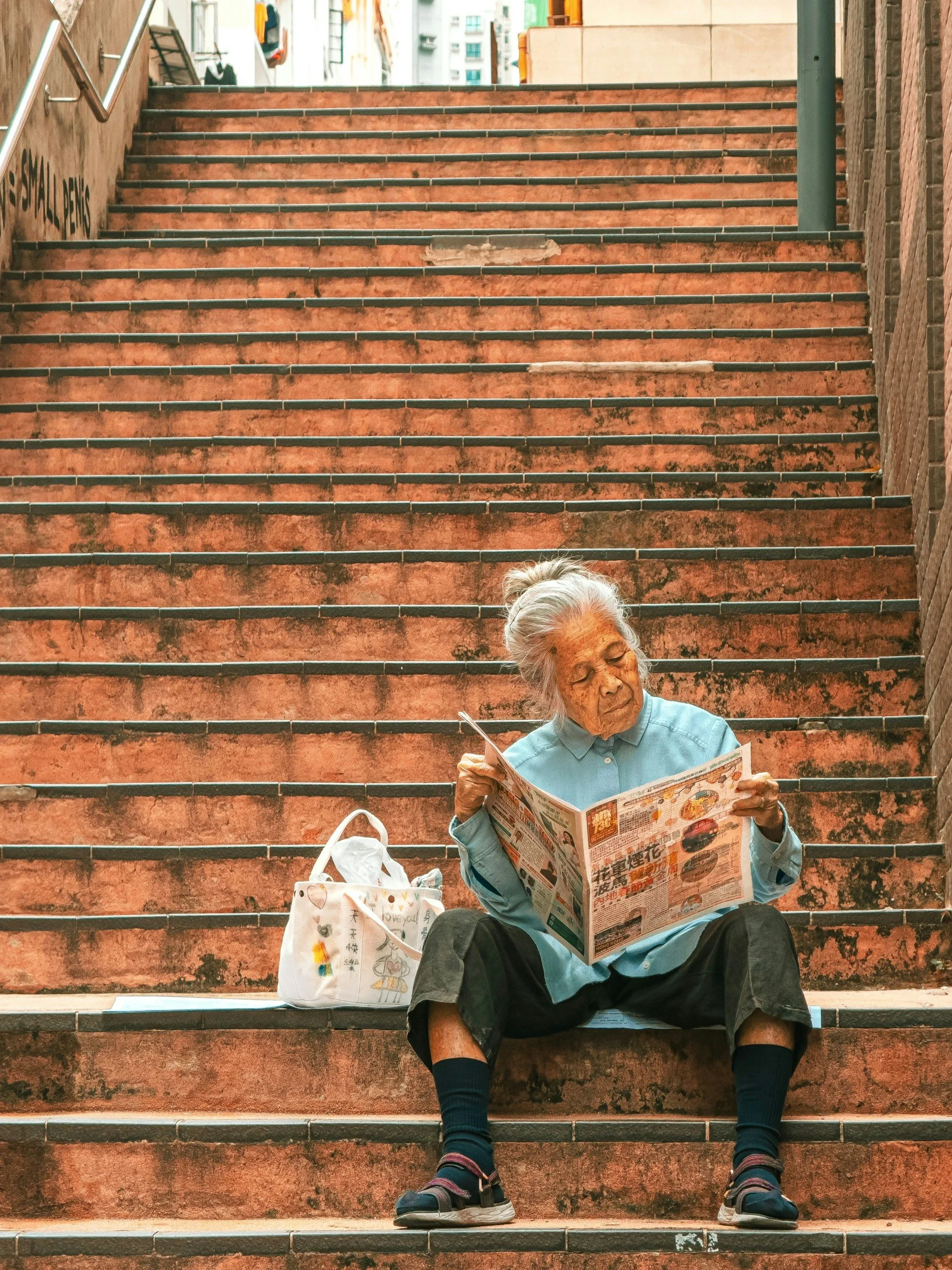 An elderly woman sitting on outdoor stairs, reading a newspaper, with a bag and a backpack beside her. She is wearing a light blue shirt, black shorts, and sneakers.