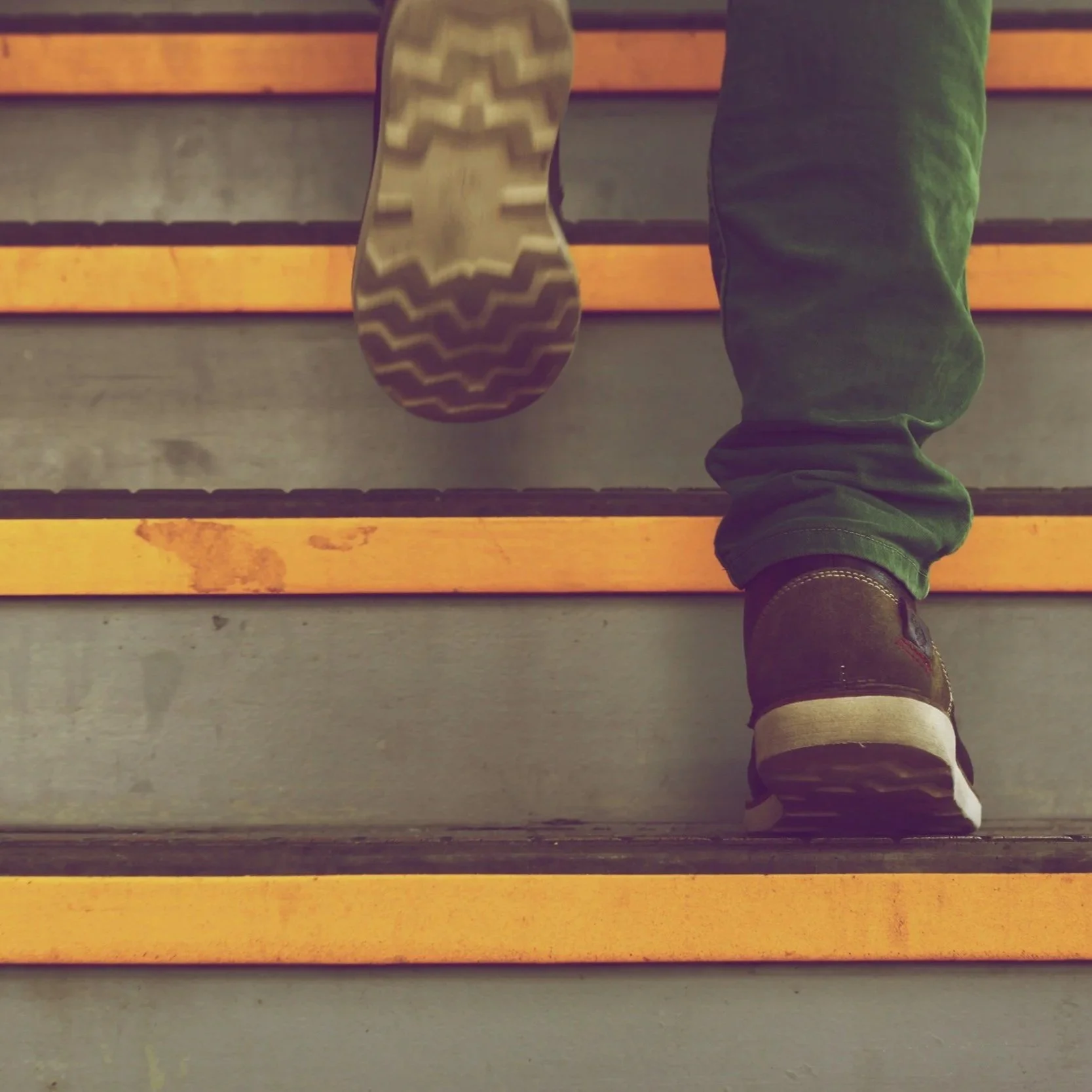 A person's feet walking up a set of wooden stairs, wearing black shoes with white soles and dark pants.