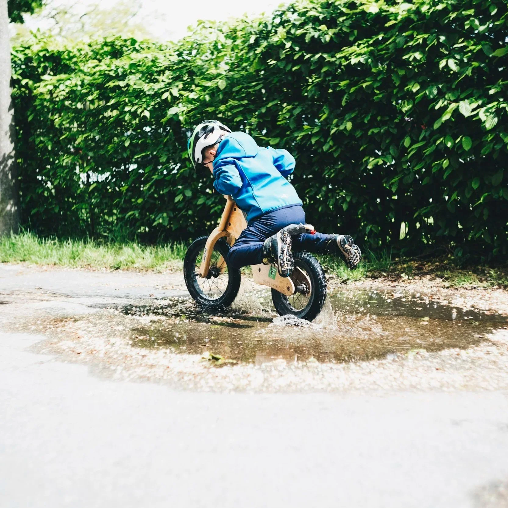 Child riding a small wooden bicycle through a puddle on a wet path, with lush green bushes in the background.