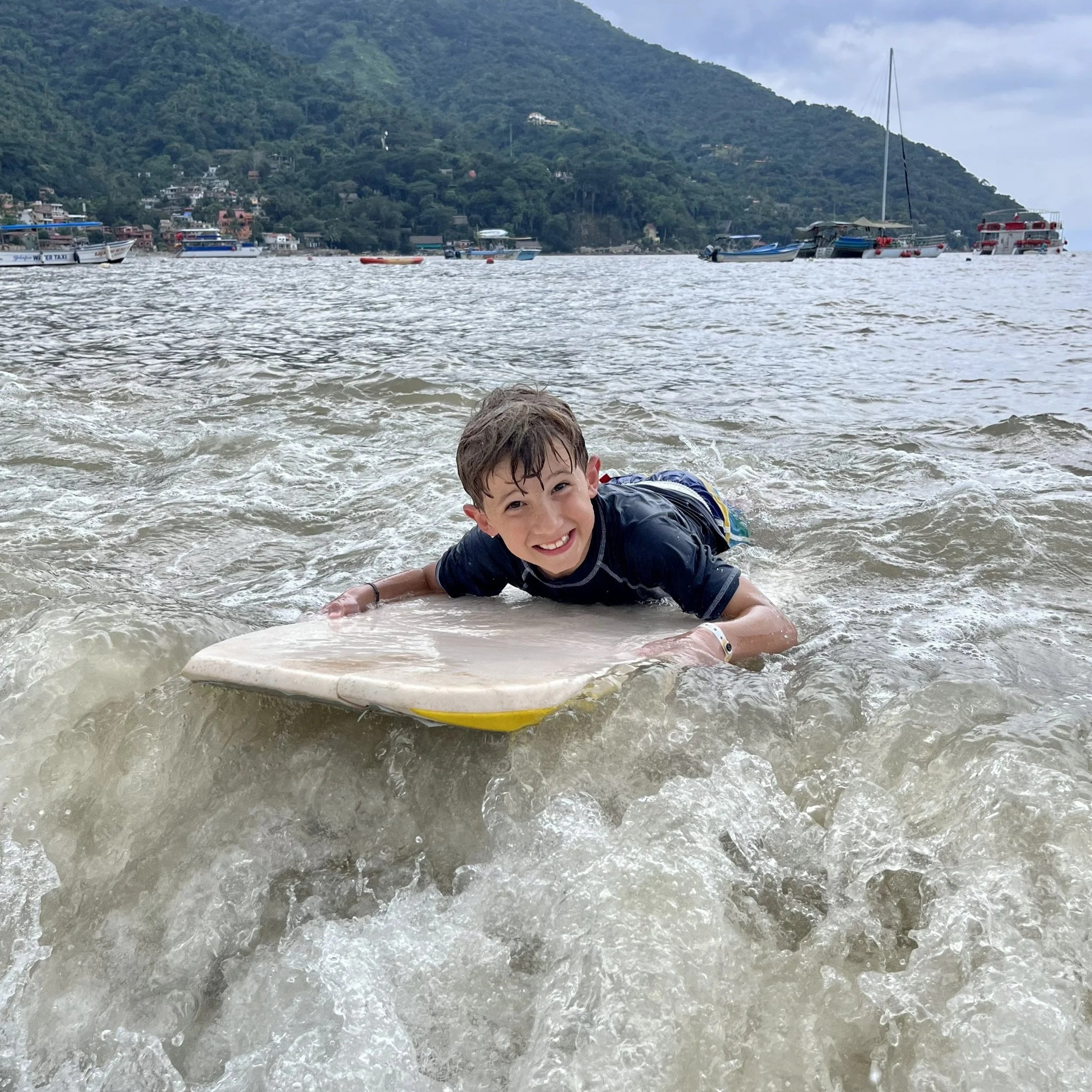 A young boy smiling while lying on a paddleboard in the water near boats and a hillside with houses.
