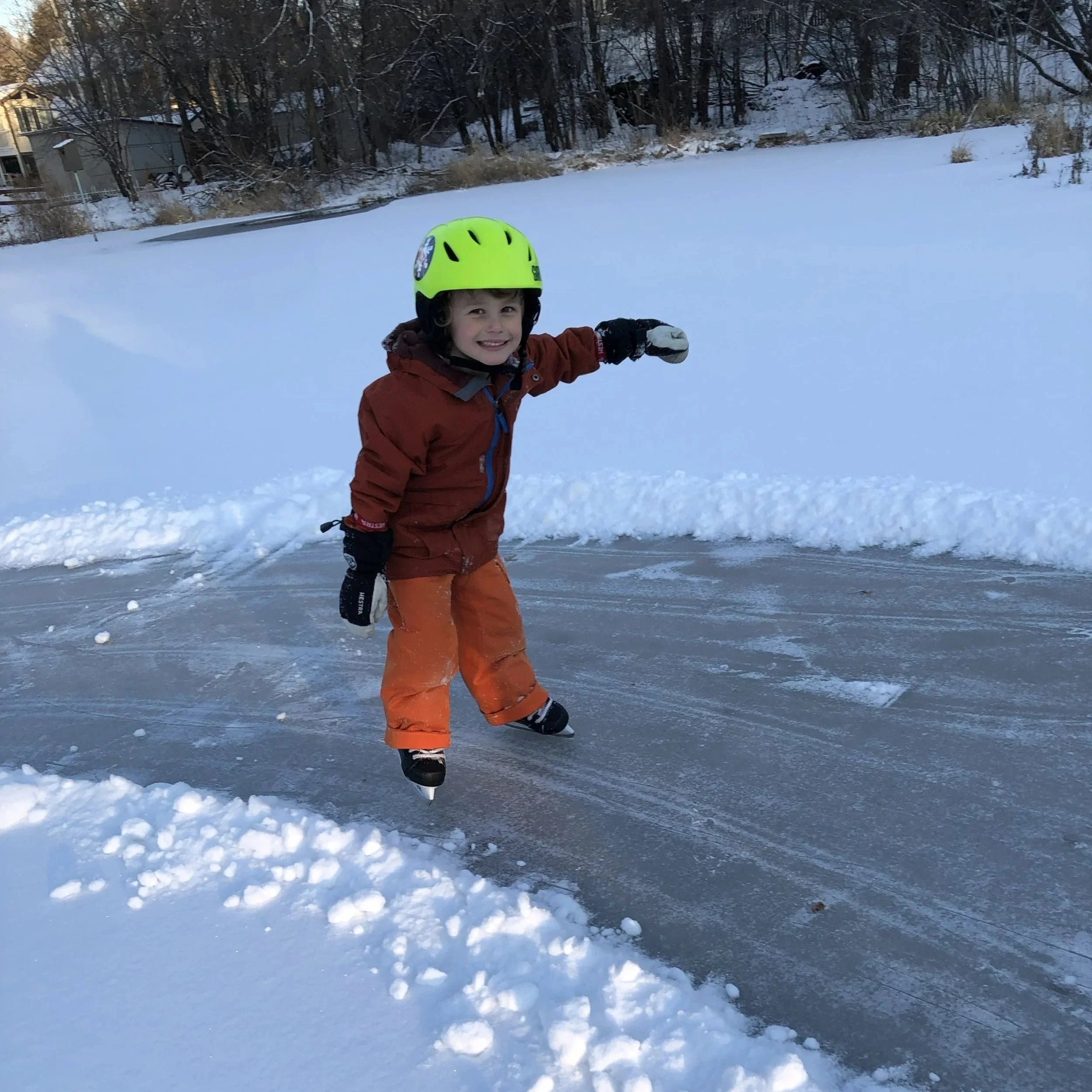 Young boy in a red winter jacket and orange snow pants wearing a bright yellow helmet, ice skating on a snow-covered outdoor surface with snow and trees in the background.