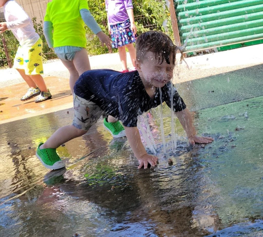 A young boy crawling on the water splash pad with water spraying from above, while other children play in the background.