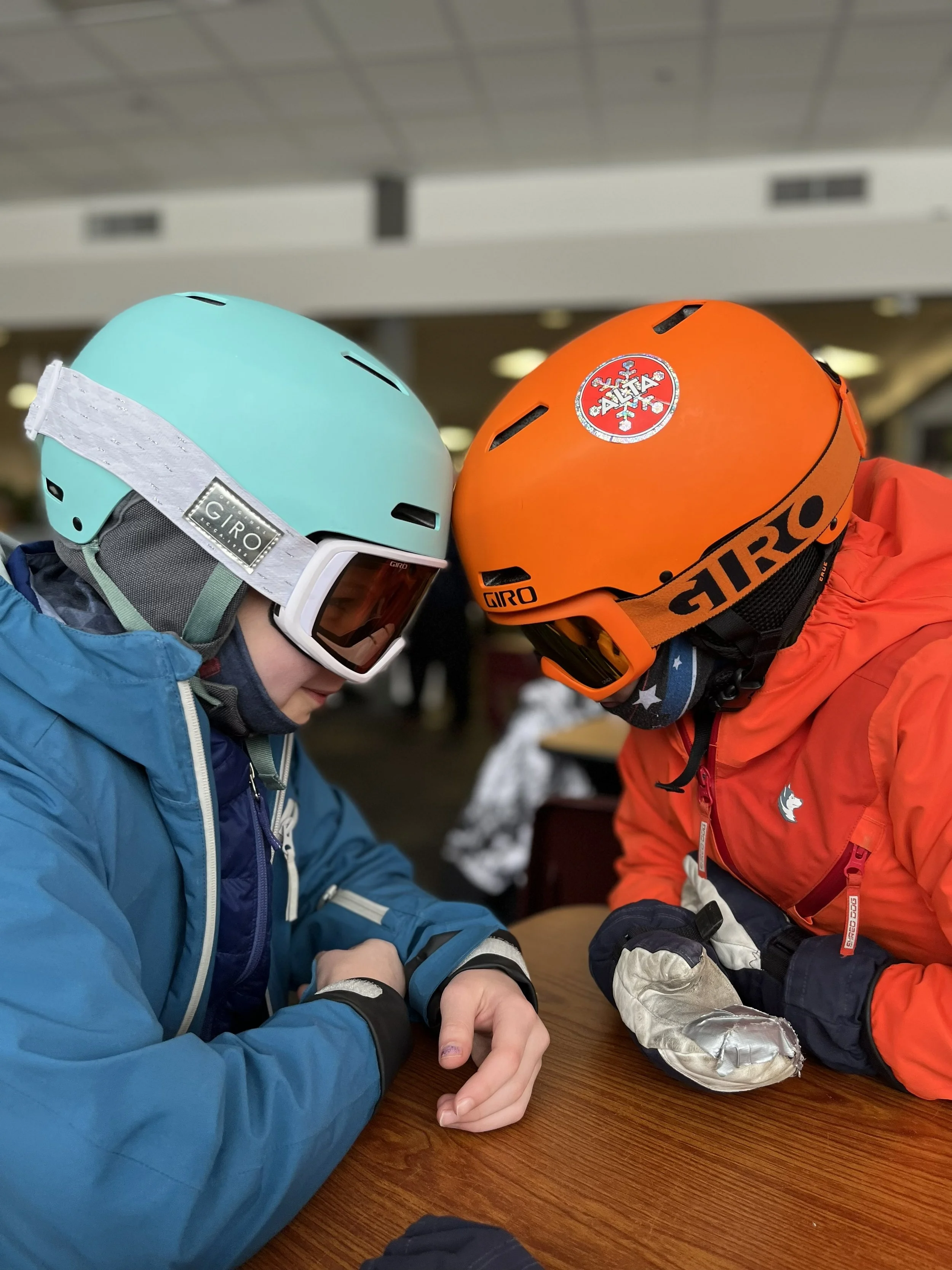 Two skiers wearing helmets, goggles, and ski jackets are sitting at a wooden table with their heads touching, possibly in a ski lodge or rest area.